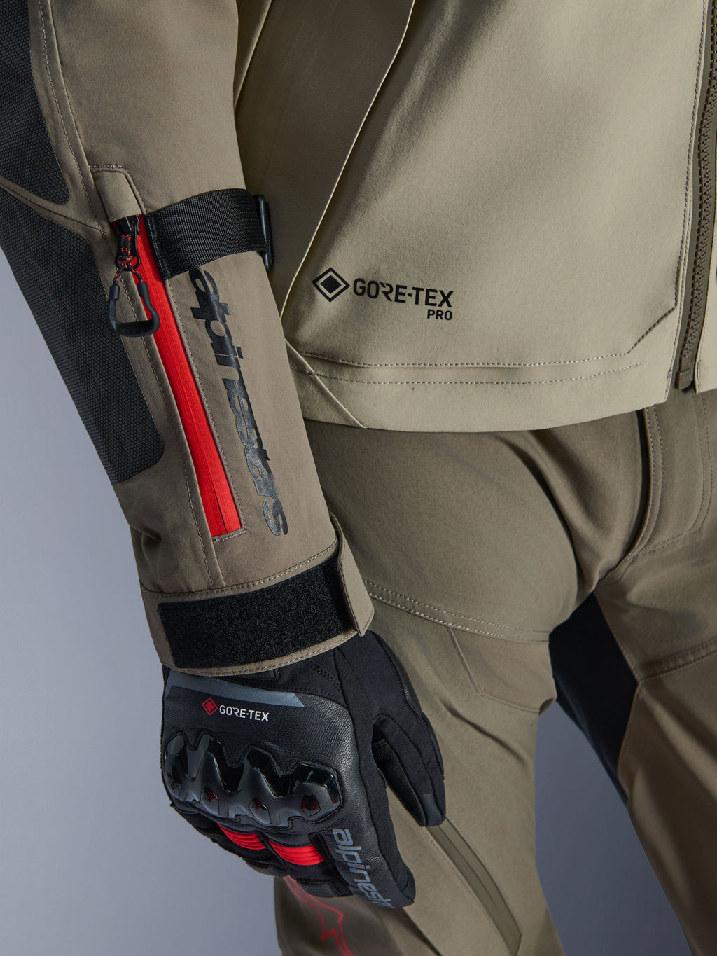 Nærbillede af Alpinestars Nazca 3L GORE-TEX PRO Jakke i Walnut Rock Brown, med røde lynlåsdetaljer på ærmet, GORE-TEX PRO-branding og kombineret med sorte GORE-TEX adventure-kørehandsker.