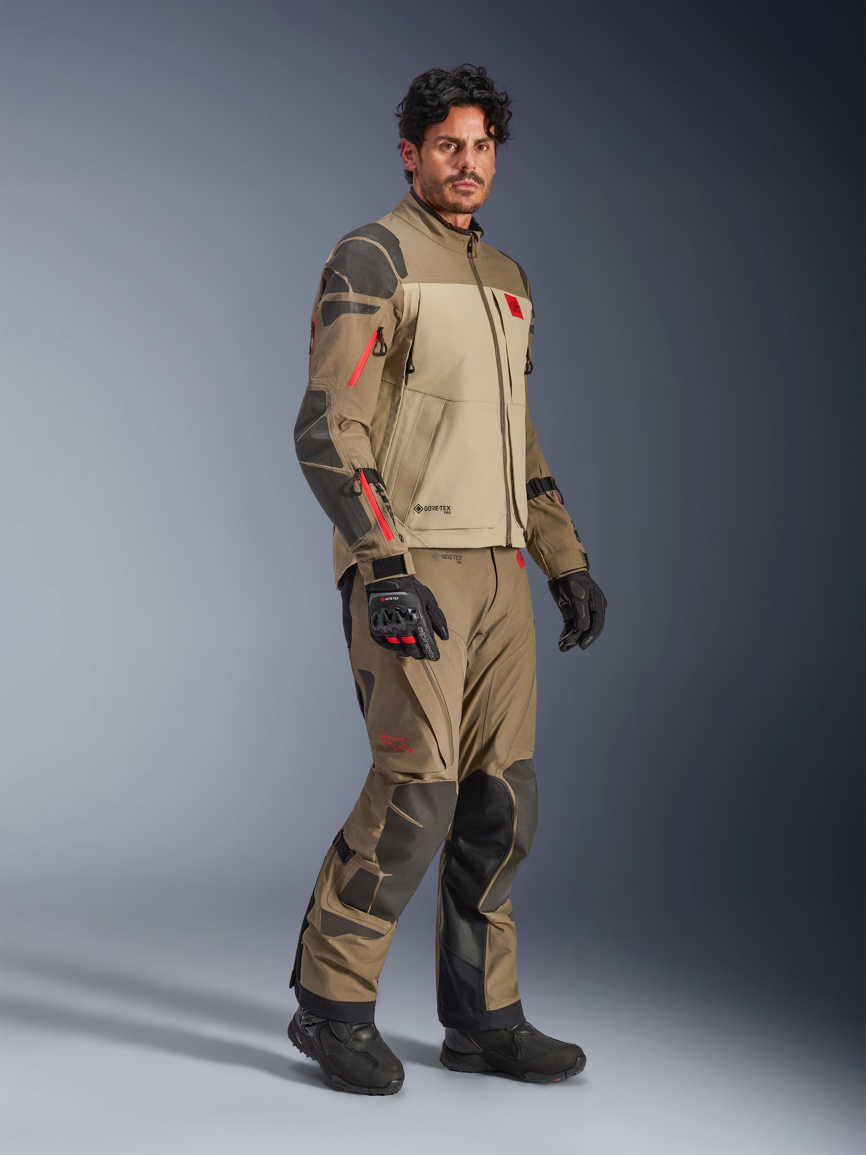 Alpinestars Nazca 3L GORE-TEX PRO jakke, Walnut Rock Brown, adventure motorcykeljakke båret med matchende bukser og sorte handsker, med mørkegrå forstærkninger, røde lynlåsdetaljer og en højtydende lamineret skal.