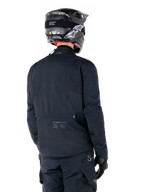 Alpinestars St-7 Prime GORE-TEX-jakke, sort, set bagfra på en rytter iført jakken med matchende hjelm og bukser, med Alpinestars-branding og geografiske koordinater på den nederste del af ryggen.