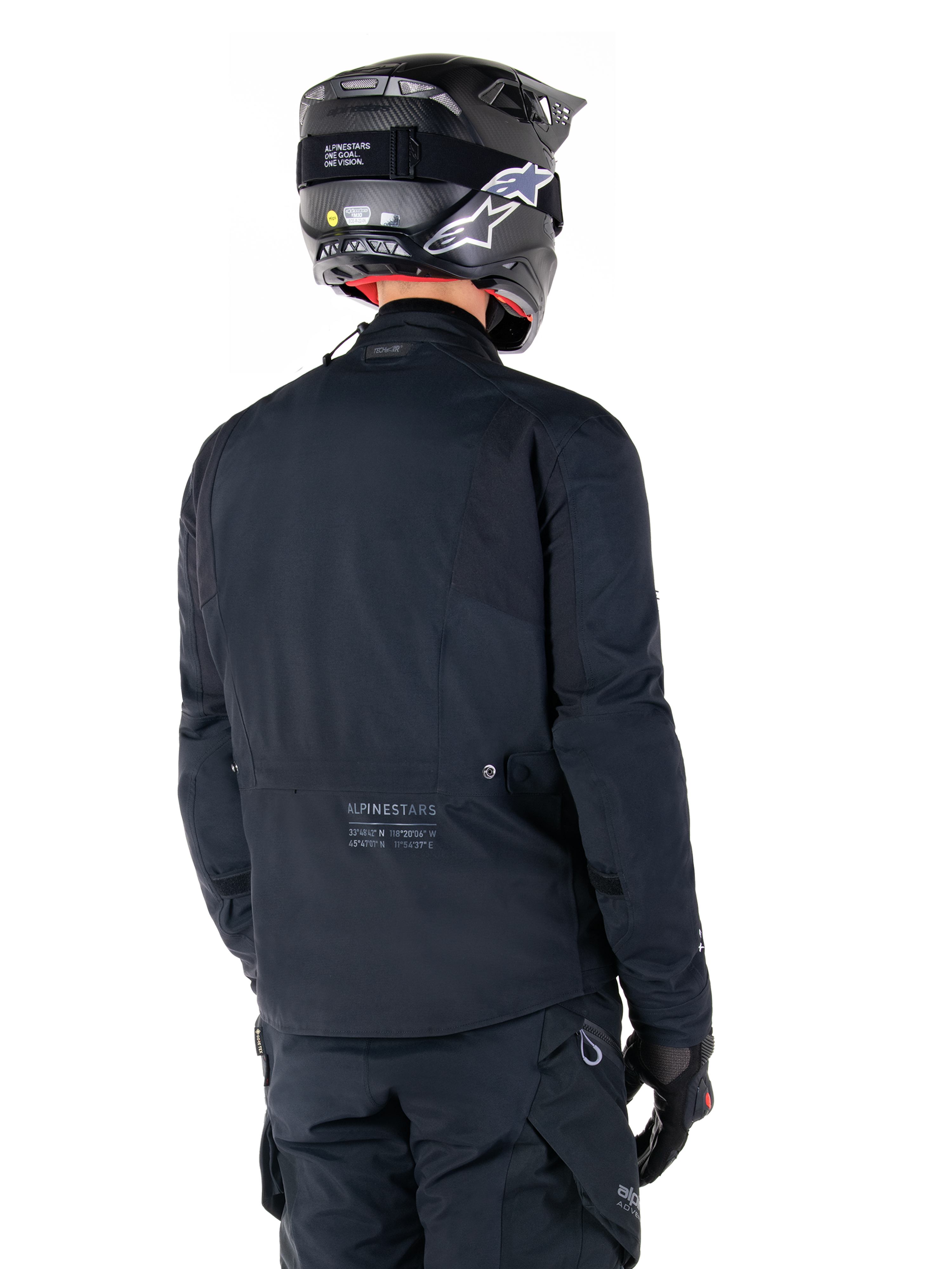 Alpinestars St-7 Prime GORE-TEX-jakke, sort, set bagfra på en rytter iført jakken med matchende hjelm og bukser, med Alpinestars-branding og geografiske koordinater på den nederste del af ryggen.