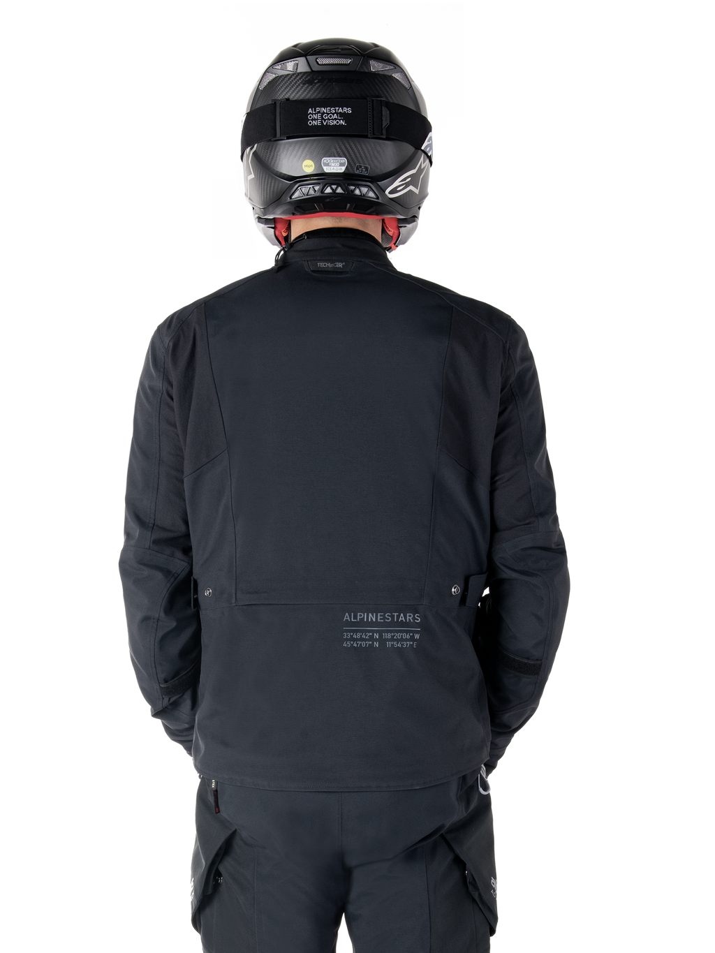 Alpinestars St-7 Prime GORE-TEX-jakke, sort, set bagfra på en rytter, med diskret logo og geografiske koordinater på den nederste del af ryggen, bæres med matchende sort hjelm og bukser, strømlinet og teknisk motorcykelbeklædning