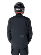 Alpinestars St-7 Prime GORE-TEX-jakke, sort, set bagfra på en rytter, med diskret logo og geografiske koordinater på den nederste del af ryggen, bæres med matchende sort hjelm og bukser, strømlinet og teknisk motorcykelbeklædning