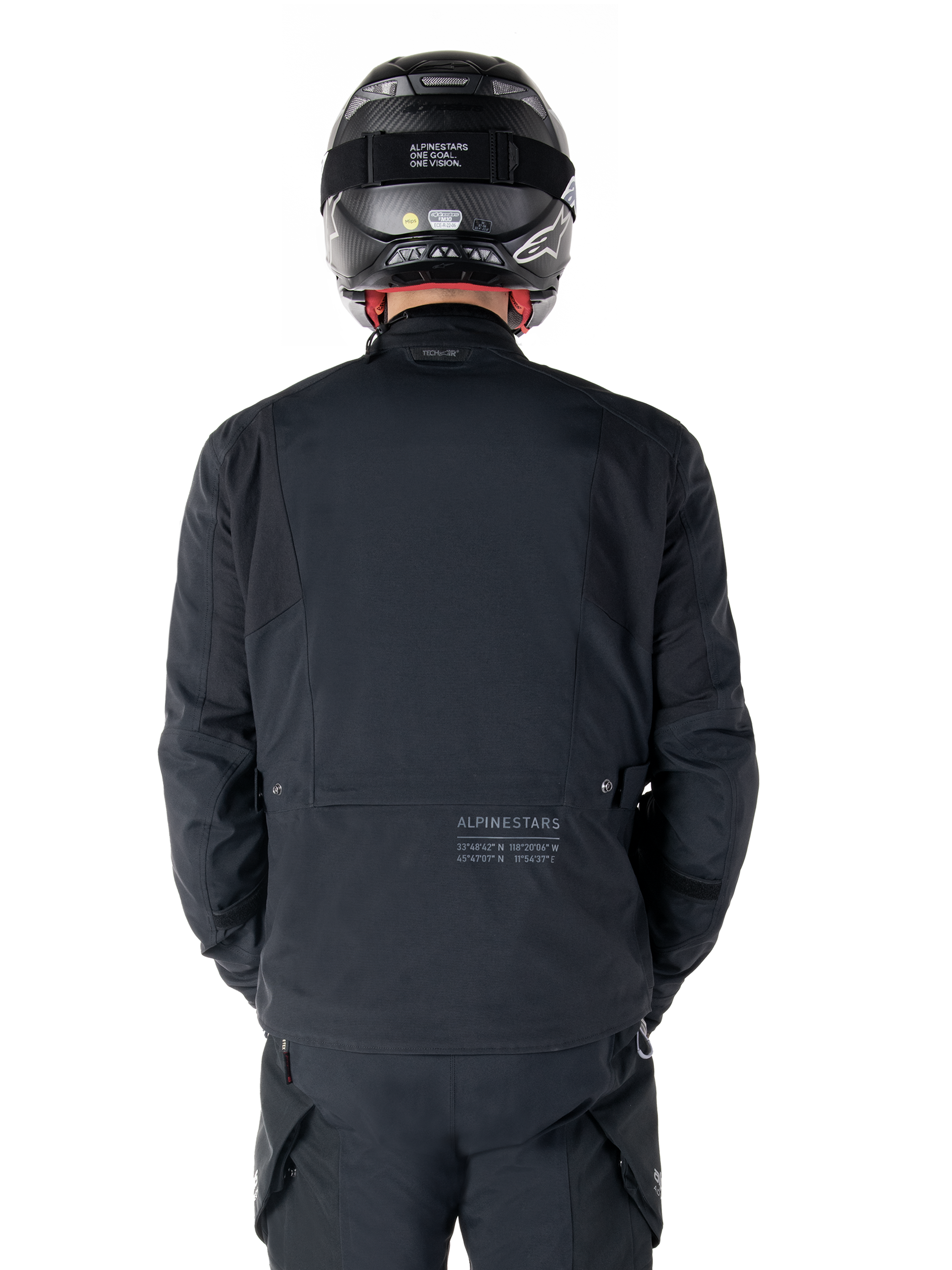 Alpinestars St-7 Prime GORE-TEX-jakke, sort, set bagfra på en rytter, med diskret logo og geografiske koordinater på den nederste del af ryggen, bæres med matchende sort hjelm og bukser, strømlinet og teknisk motorcykelbeklædning