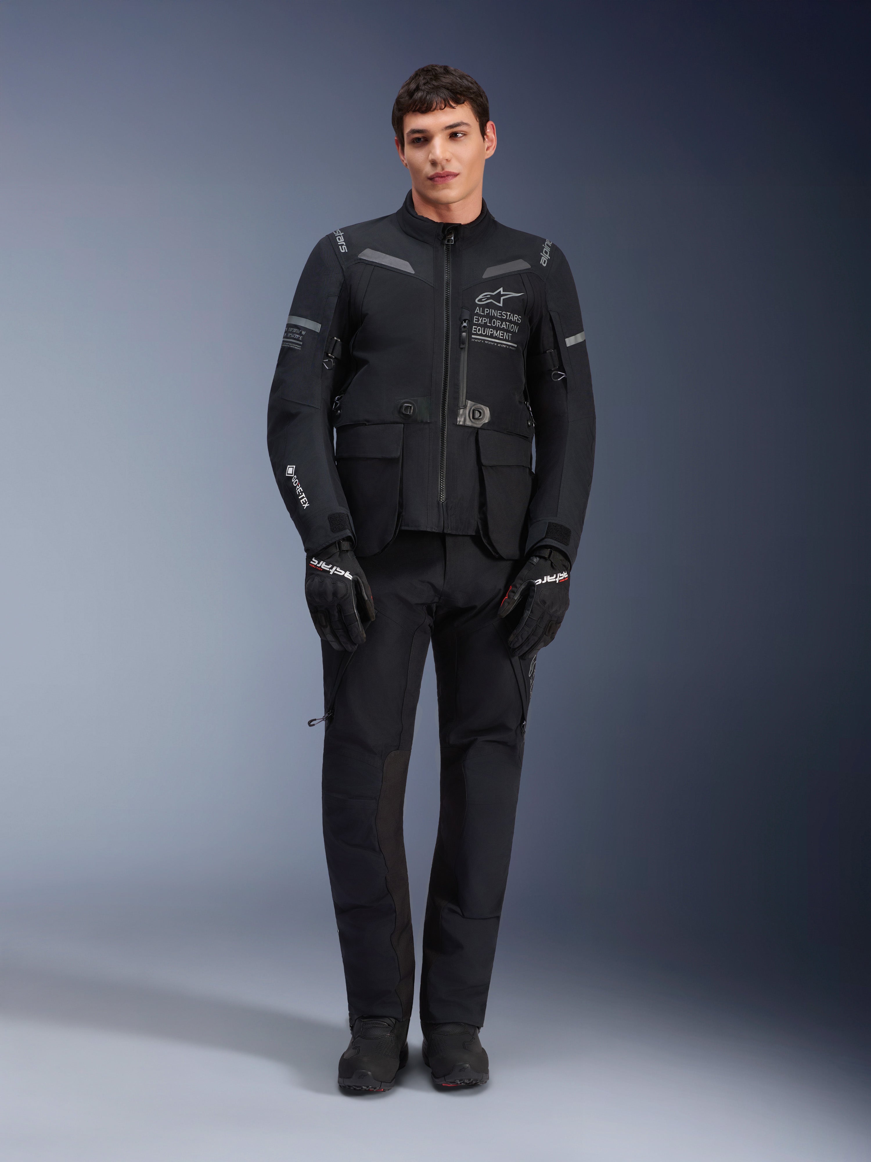 Alpinestars ST-7 2L GORE-TEX Jacket, adventure touring motorcycle jacket, Black and Dark Gray, worn with matching black riding pants and gloves, featuring multiple cargo pockets, reflective accents, and Alpinestars Exploration Equipment chest branding, functional design for road and off-road riding