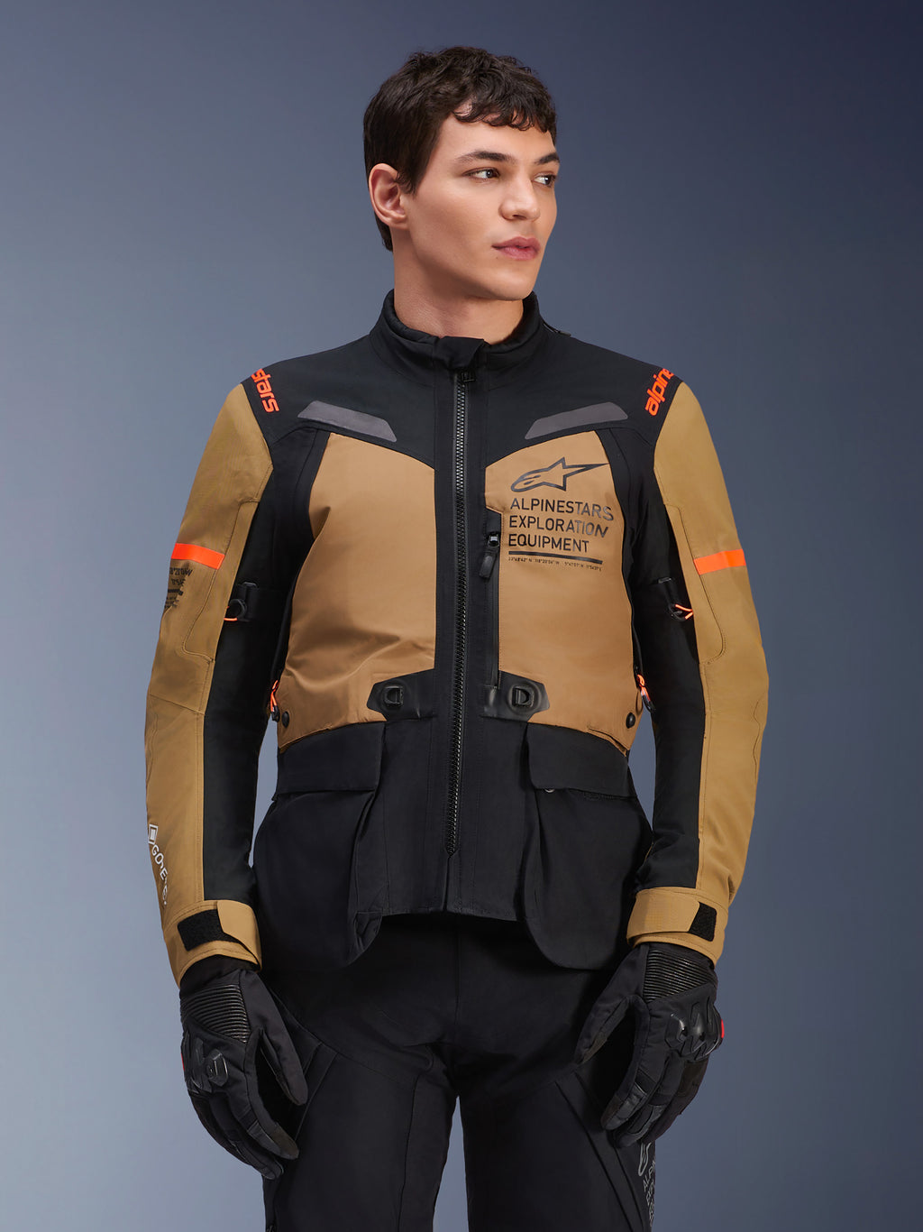 Alpinestars ST-7 2L GORE-TEX jakke, motorcykeljakke til adventure touring, Gobi Brown og sort med orange detaljer, båret af en mandlig model, med Exploration Equipment-branding og GORE-TEX-teknologi, slidstærkt vandtæt køreudstyr