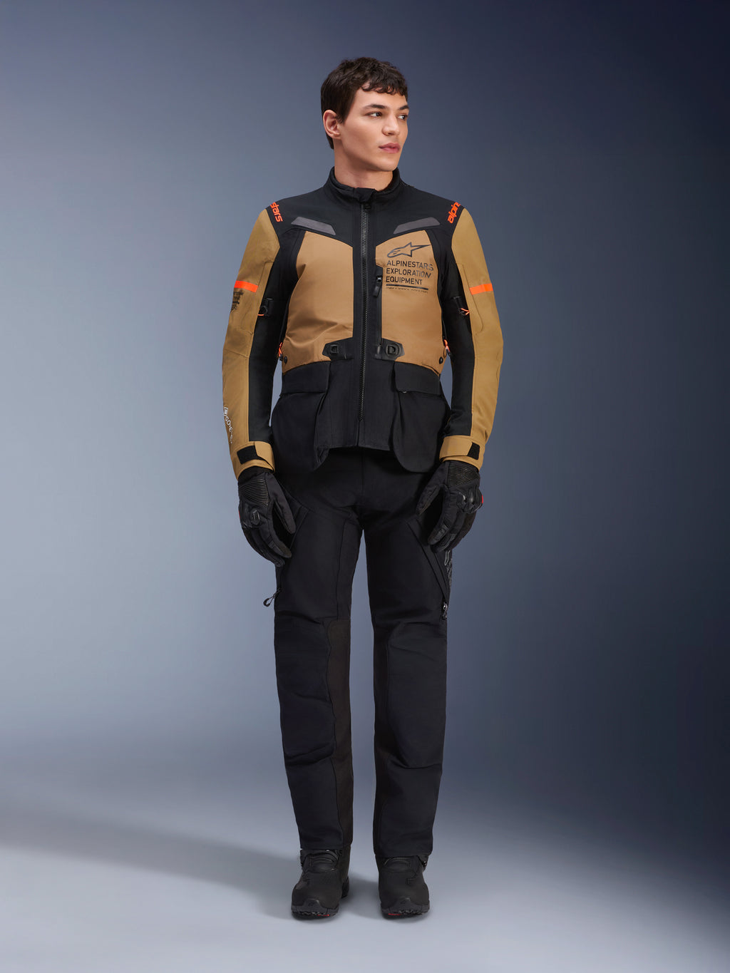 Mandlig model iført Alpinestars ST-7 2L GORE-TEX-jakke i Gobi Brown og sort, adventure-motorcykeludstyr med matchende sorte bukser og handsker, med Alpinestars Exploration Equipment-branding på brystet, funktionelt vandtæt design til touring på vej og off-road