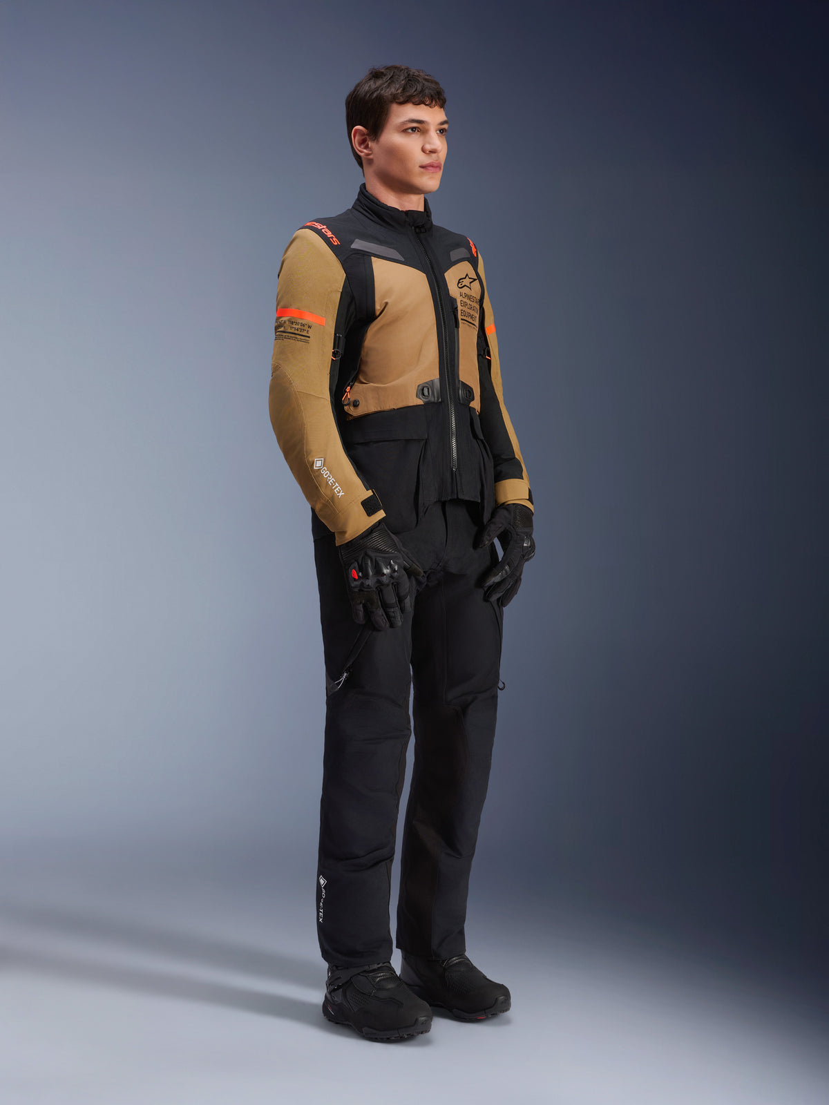 Alpinestars ST-7 2L GORE-TEX-jakke, motorcykeljakke, Gobi Brun og Sort, båret af en model med matchende sorte bukser og handsker, med brune paneler på brystet og ærmerne, sorte tekniske detaljer og orange branding, adventure-touring motorcykeludstyr.