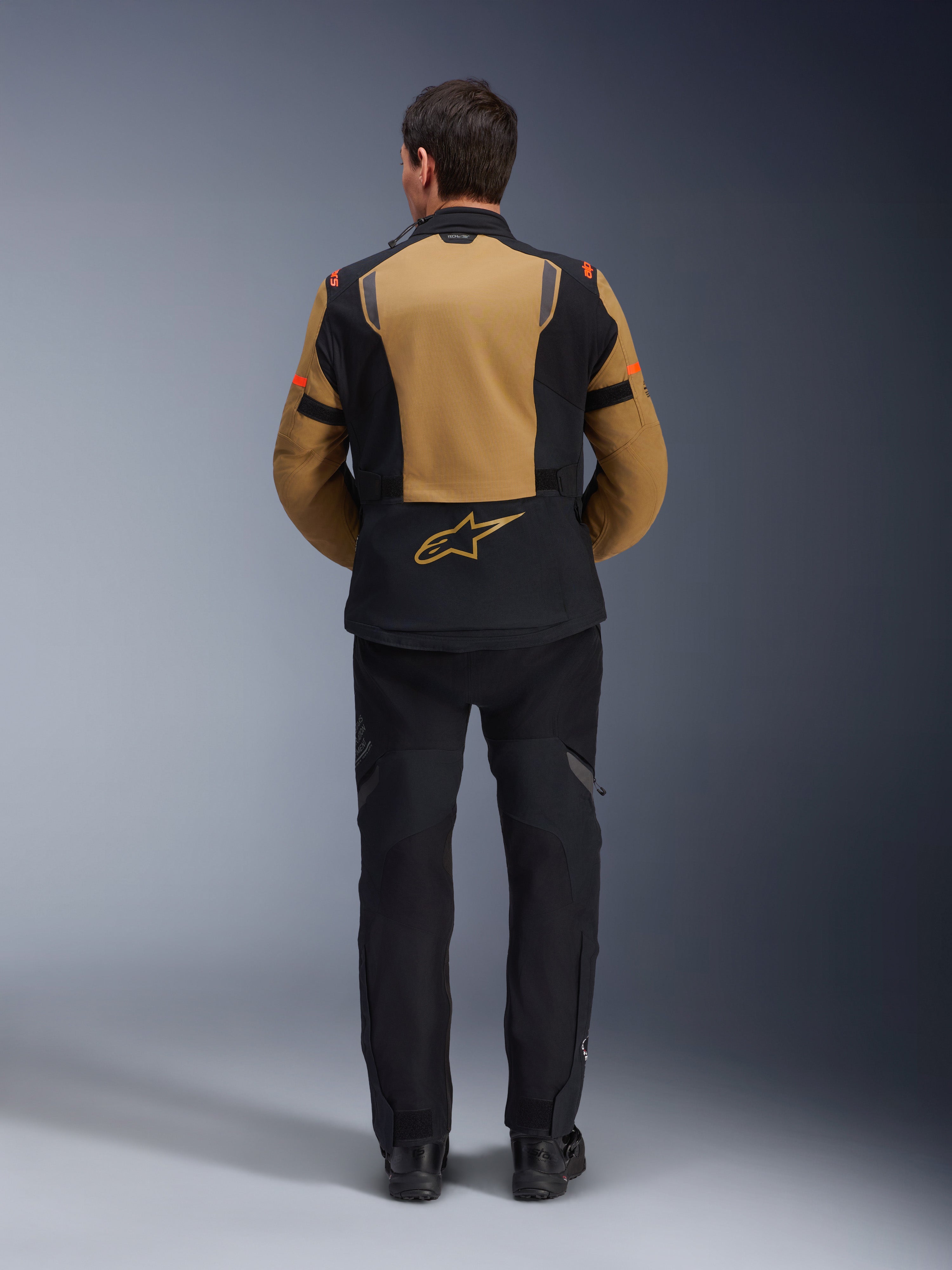 Alpinestars ST-7 2L GORE-TEX jakke, adventure motorcykeljakke, Gobi Brown og sort, set bagfra der viser ergonomiske paneler og Alpinestars-logo, båret med sorte kørebukser og støvler, vandtæt touringudstyr
