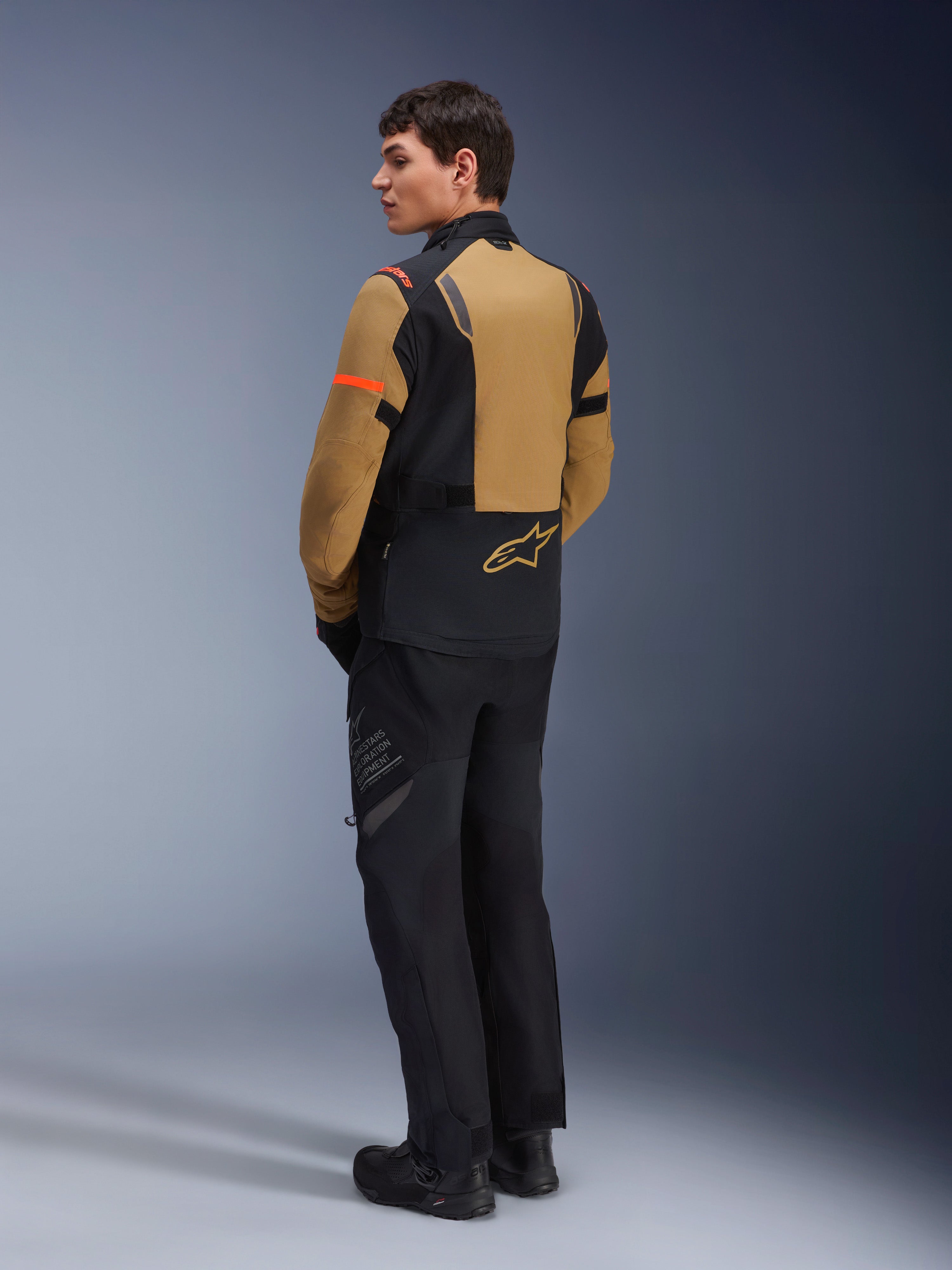 Alpinestars ST-7 2L GORE-TEX jakke, adventure-motorcykeljakke, Gobi Brown og sort, set bagfra på en model iført matchende kørebukser og støvler, med orange detaljer på ærmerne, vandtæt lamineret yderskal og Alpinestars-logo, designet til touring og landevejskørsel