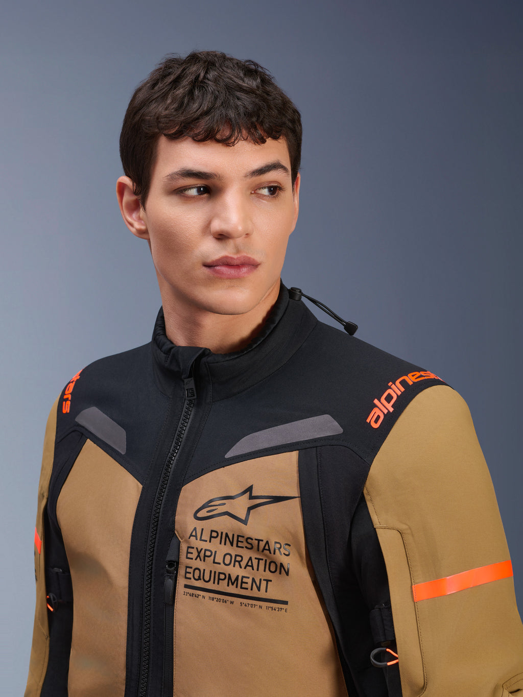 Alpinestars ST-7 2L GORE-TEX-jakke, Gobi Brown og sort, vist på en mandlig model i et studiemiljø, med tydelig 'ALPINESTARS EXPLORATION EQUIPMENT'-branding på brystet, orange skulderlogoer og teknisk vandtæt tekstilkonstruktion til adventure-kørsel.