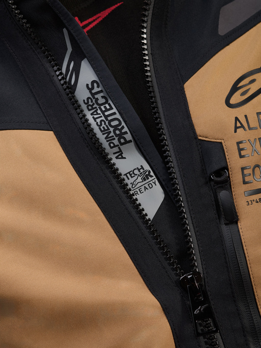 Nærbillede af Alpinestars ST-7 2L GORE-TEX-jakke i Gobi Brown og sort, med 'ALPINESTARS PROTECTS' og 'TECH-AIR READY' branding indvendigt ved hovedlynlåsen, robust tekstilkonstruktion og brystlogoer
