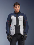 Alpinestars ST-7 2L GORE-TEX jakke, adventure touring motorcykeljakke, isgrå og mørkeblå med røde detaljer, vandtæt og åndbar, båret af en model med sorte handsker, med Alpinestars Exploration Equipment-branding på brystet