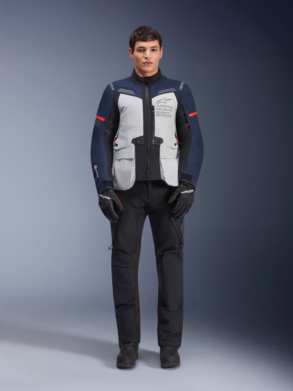 Alpinestars ST-7 2L GORE-TEX jakke, adventure touring motorcykeljakke, Ice Gray, mørkeblå og sort, båret med sorte bukser og handsker, med store cargolommer foran, røde detaljer på ærmerne og en lamineret GORE-TEX-skal for vandtæt ydeevne.
