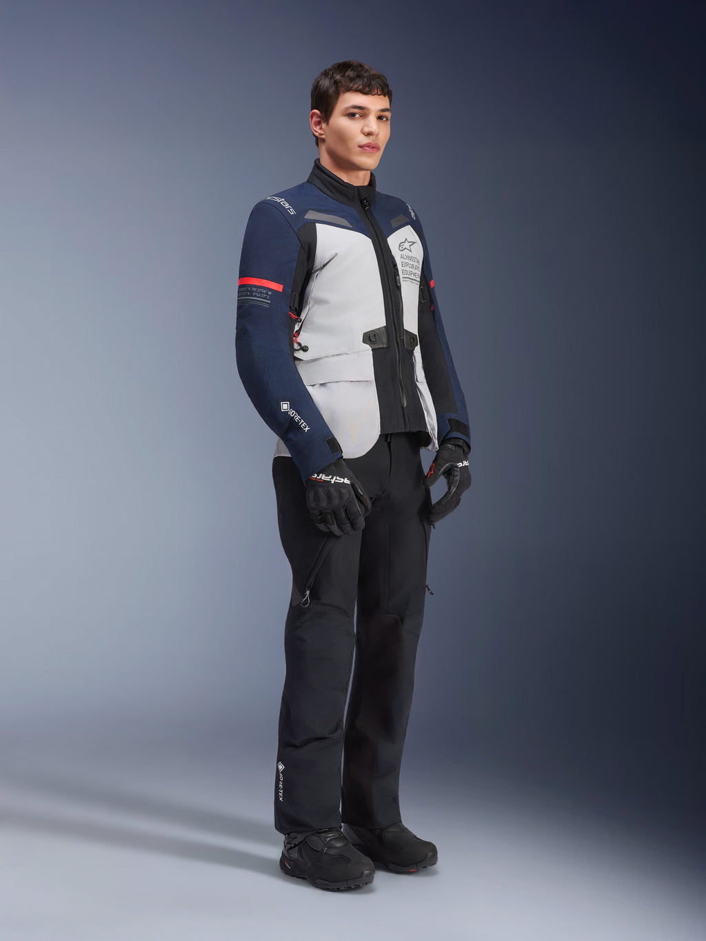 Alpinestars ST-7 2L GORE-TEX-jakke i farvekombinationen Ice Gray, Dark Blue og sort med røde detaljer, båret af en mandlig model med sorte motorcykelbukser og handsker, professionelt adventure touring-motorcykeludstyr
