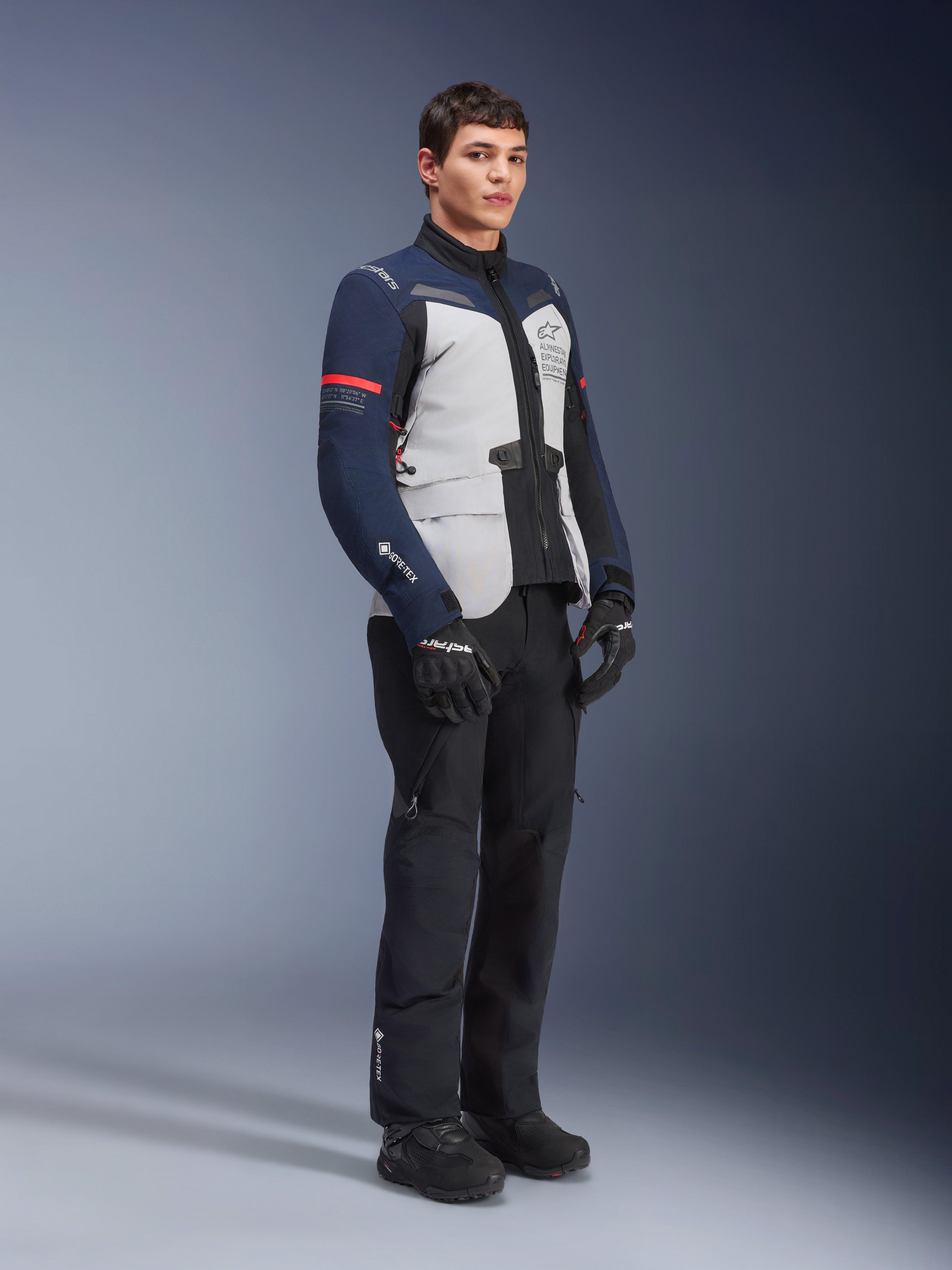 Alpinestars ST-7 2L GORE-TEX-jakke i farvekombinationen Ice Gray, Dark Blue og sort med røde detaljer, båret af en mandlig model med sorte motorcykelbukser og handsker, professionelt adventure touring-motorcykeludstyr