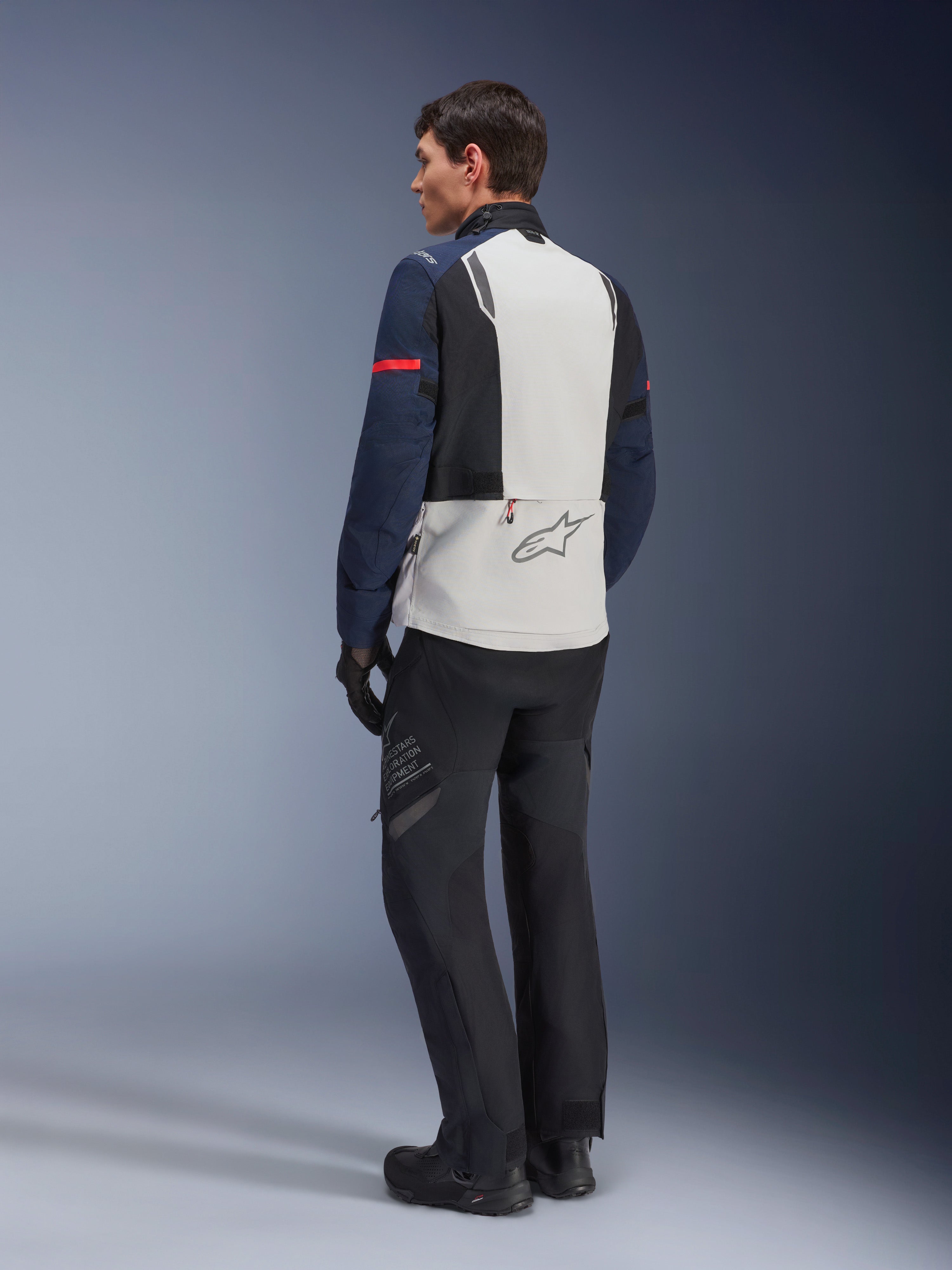 Model iført Alpinestars ST-7 2L GORE-TEX jakke i Ice Gray, Dark Blue og sort, set bagfra, lysegråt rygpanel med Alpinestars-logo, mørkeblå ærmer med rød stribe, sorte adventure-kørebukser og -støvler, motorcykel touring-udstyr