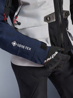 Alpinestars ST-7 2L GORE-TEX jakke, nærbillede af mørkeblåt ærme med hvidt GORE-TEX-logo, isgrå torso med sorte detaljer og røde snoreaccenter, båret med sorte Alpinestars-handsker og -bukser, adventure touring-motorcykeludstyr