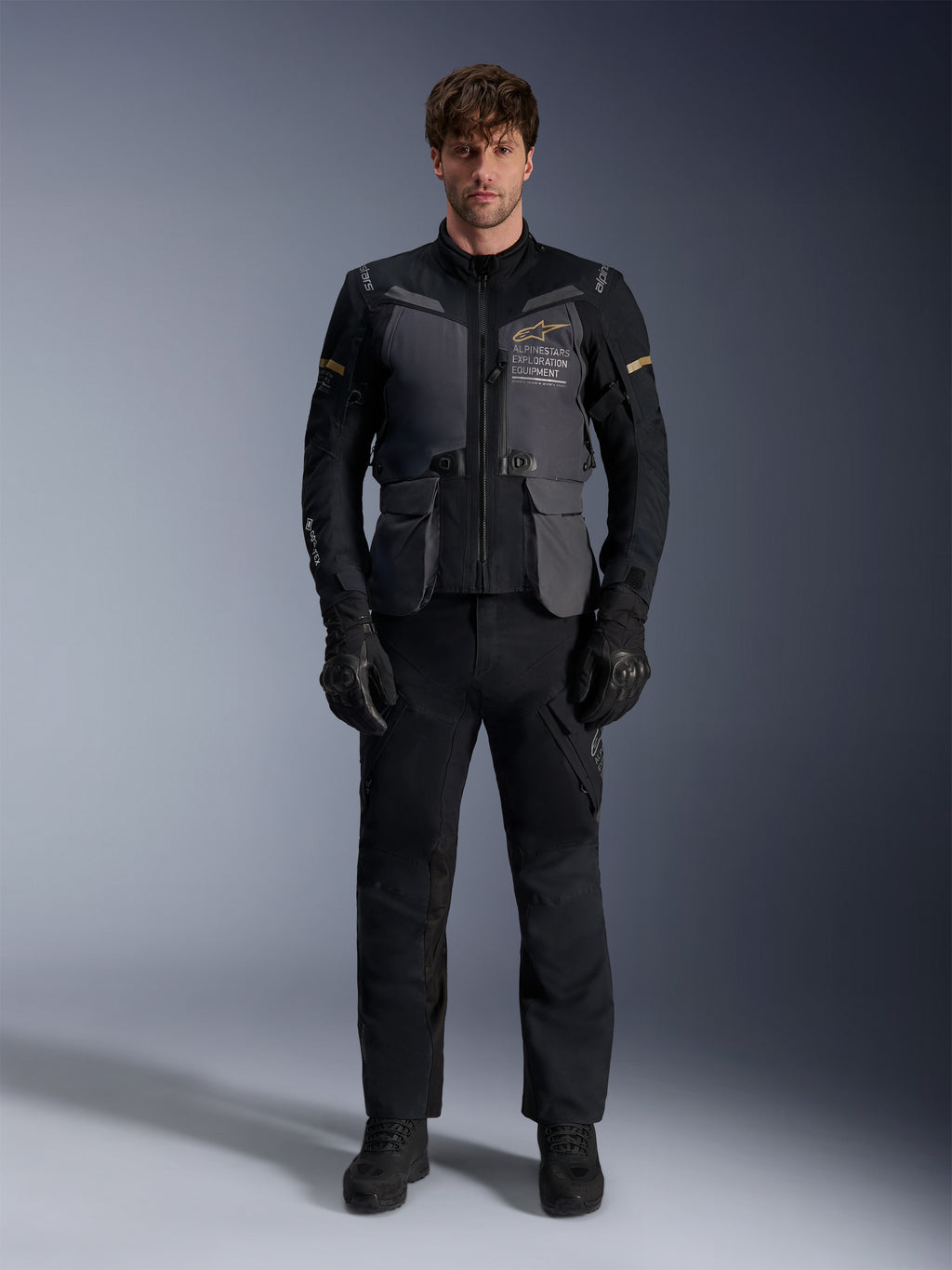 Mandlig model iført Alpinestars ST-7 2L GORE-TEX-jakke i Deep Gray Black Dark Khaki, med 'ALPINESTARS EXPLORATION EQUIPMENT'-branding på brystet, kombineret med matchende sorte kørebukser, handsker og støvler, studiebillede set forfra.