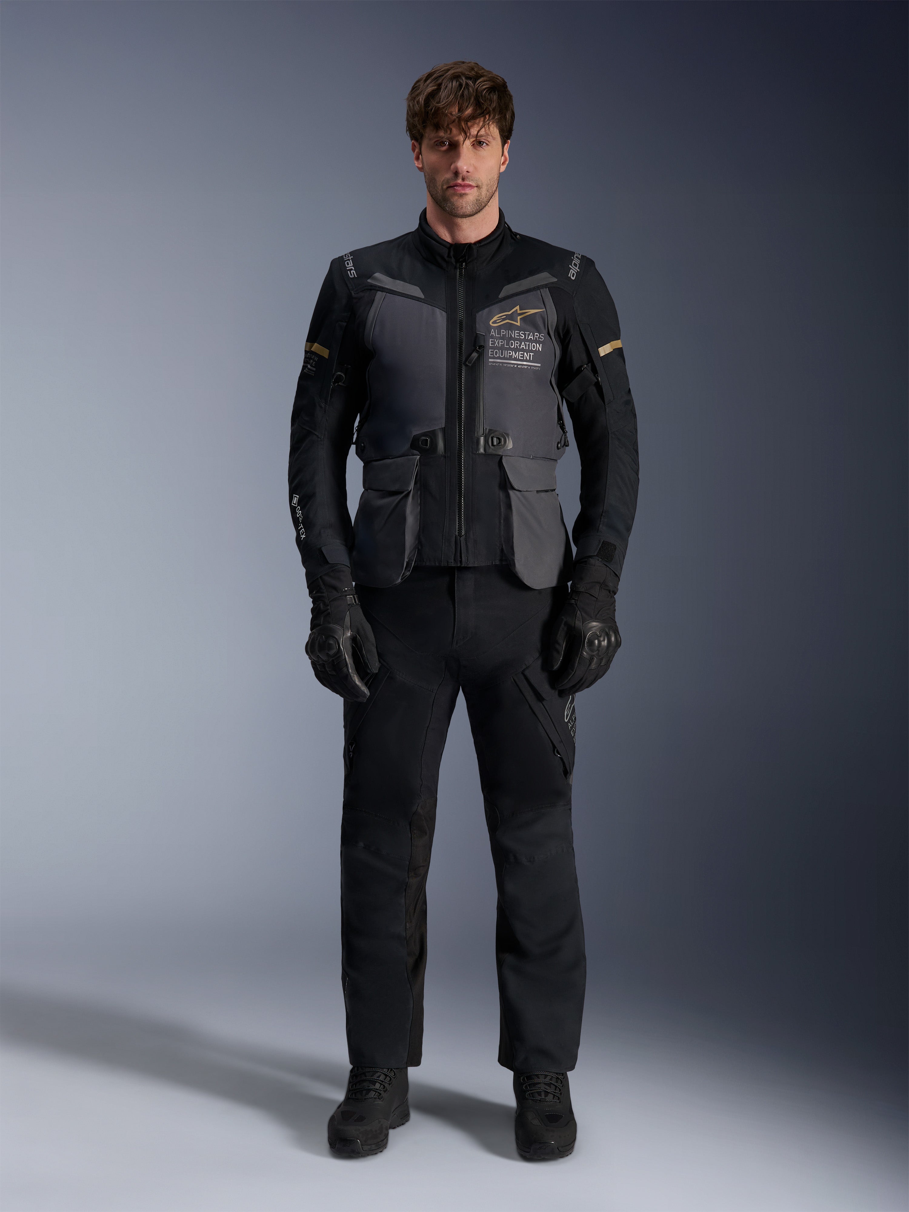 Mandlig model iført Alpinestars ST-7 2L GORE-TEX-jakke i Deep Gray Black Dark Khaki, med 'ALPINESTARS EXPLORATION EQUIPMENT'-branding på brystet, kombineret med matchende sorte kørebukser, handsker og støvler, studiebillede set forfra.