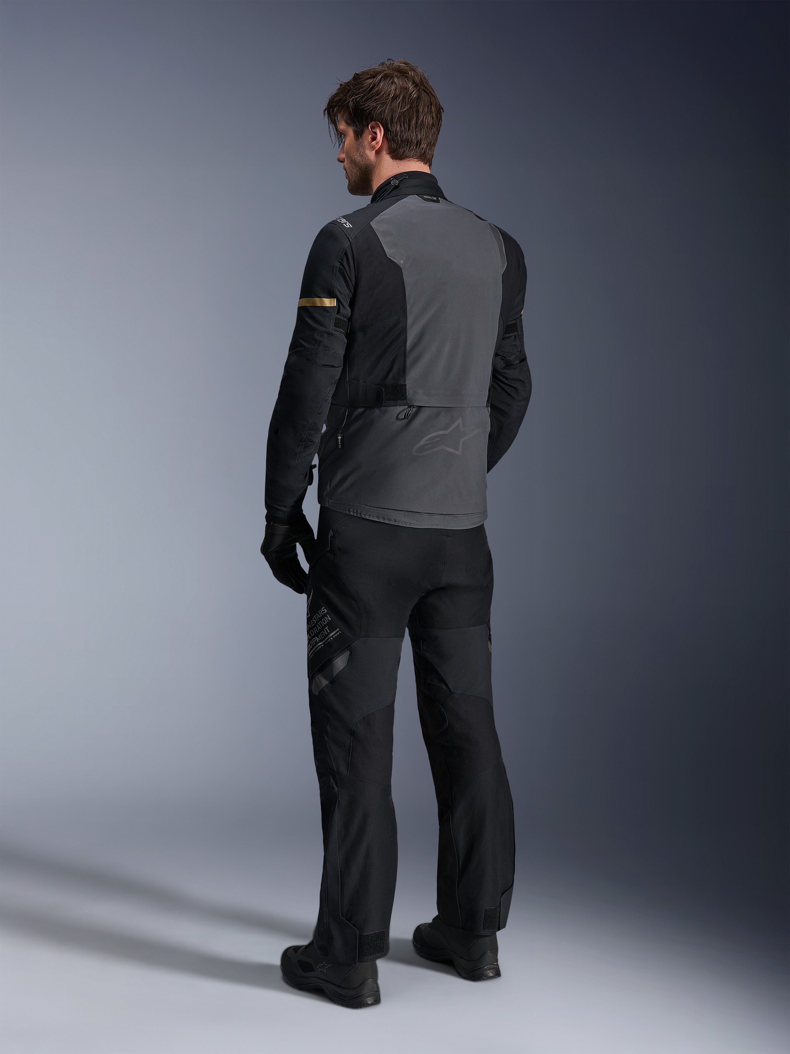 Alpinestars ST-7 2L GORE-TEX-jakke, adventure touring-udstyr, mørkegrå og sort med mørke khaki-detaljer, set bagfra af en kører iført jakken og matchende motorcykelbukser, vandtæt lamineret konstruktion
