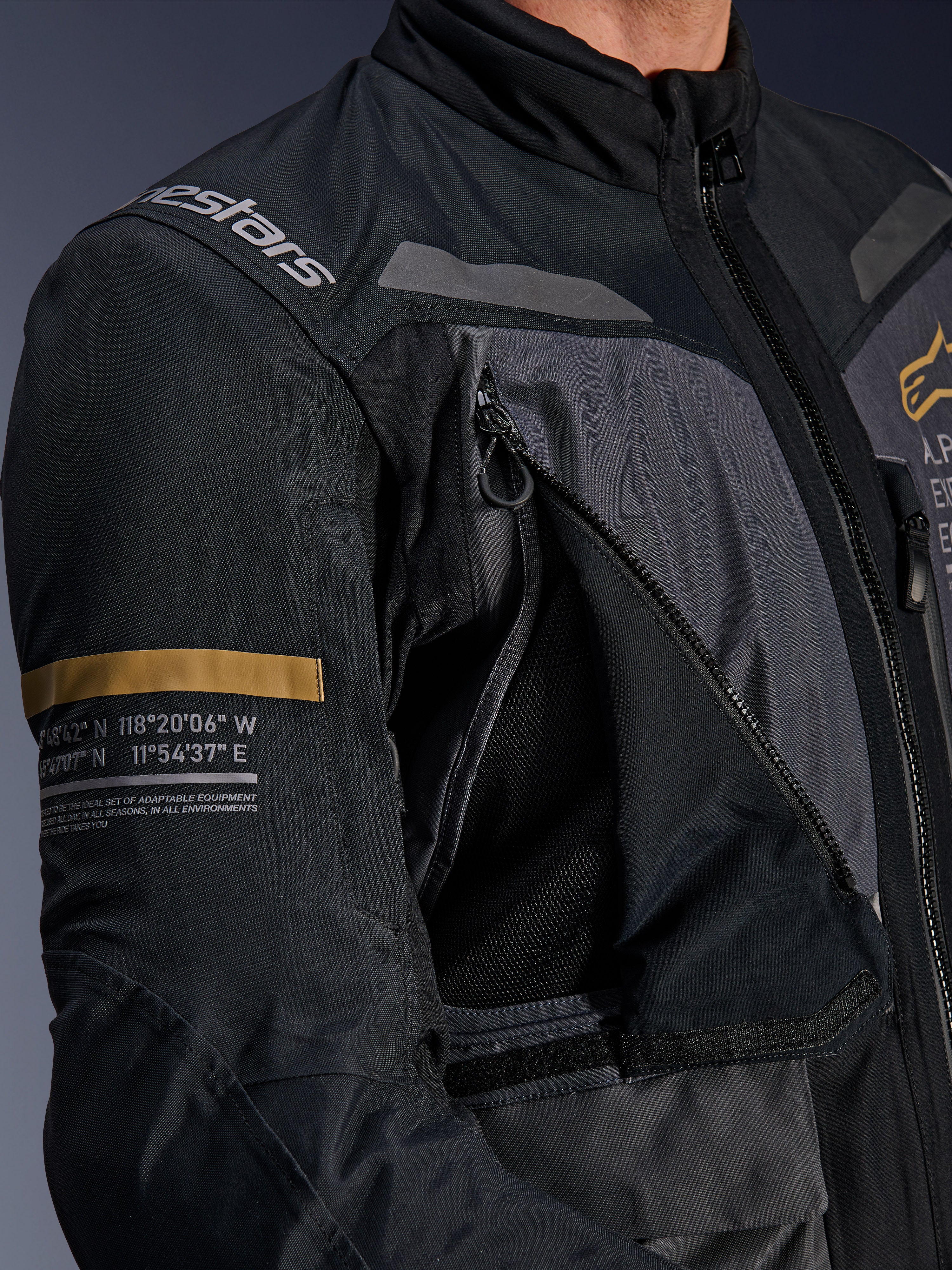 Alpinestars ST-7 2L GORE-TEX jakke, mørkegrå og sort, nærbillede af åbent ventilationspanel på brystet med lynlås og mesh, Alpinestars-logo på skulderen, khaki-stribe på ærmet med trykte geografiske koordinater, teknisk adventure-touring motorcykeludstyr