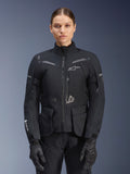 Kvinde iført Alpinestars Stella ST-7 2L GORE-TEX-jakke, sort med mørkegrå detaljer, motorcykel-touringjakke med store cargolommer og høj krave, kombineret med sorte handsker og bukser, vandtæt adventure-køreudstyr
