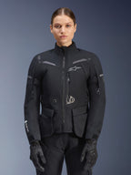 Kvinde iført Alpinestars Stella ST-7 2L GORE-TEX-jakke, sort med mørkegrå detaljer, motorcykel-touringjakke med store cargolommer og høj krave, kombineret med sorte handsker og bukser, vandtæt adventure-køreudstyr