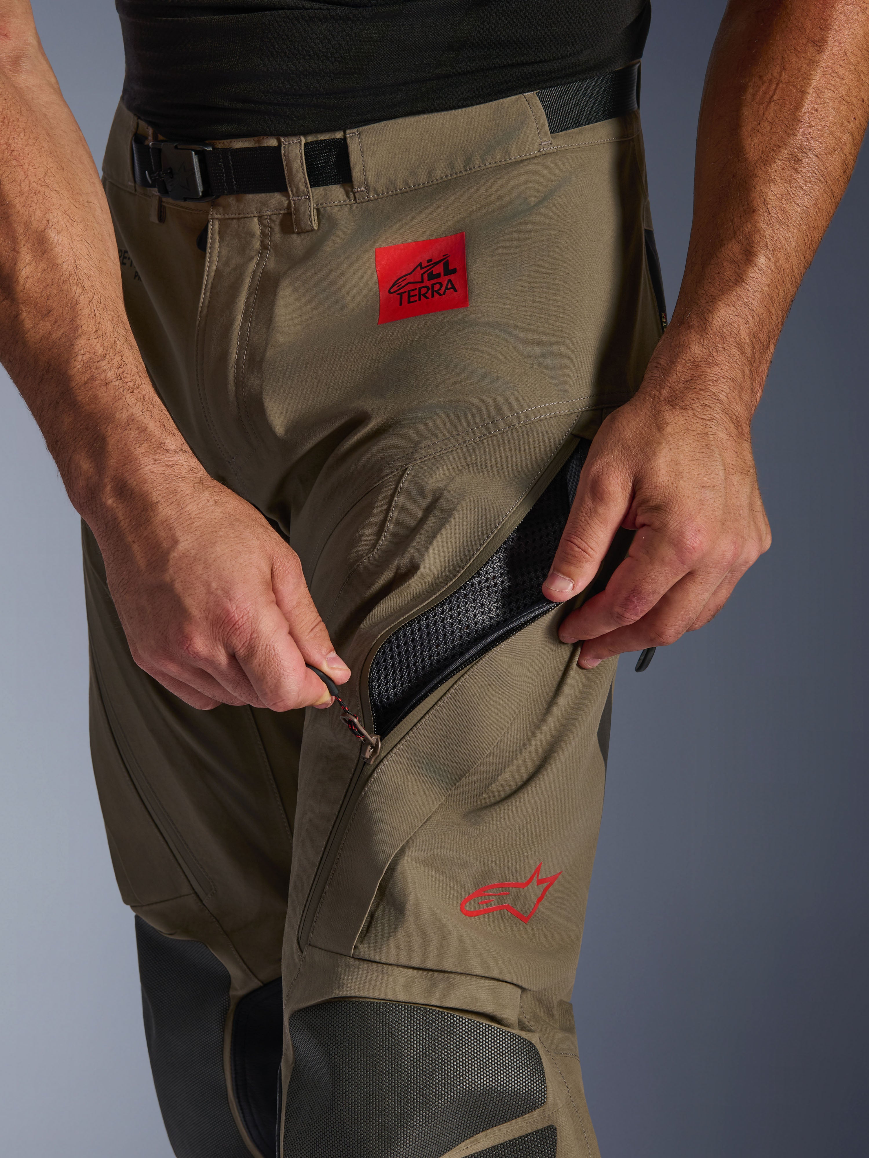 Alpinestars Nazca 3L GORE-TEX PRO bukser i Walnut Black, nærbillede af lårområdet, der viser en hånd, som åbner det store ventilationsindtag med lynlås, med et rødt ALL TERRA-mærke og rødt Alpinestars-logo, teknisk adventure-motorcykeludstyr.