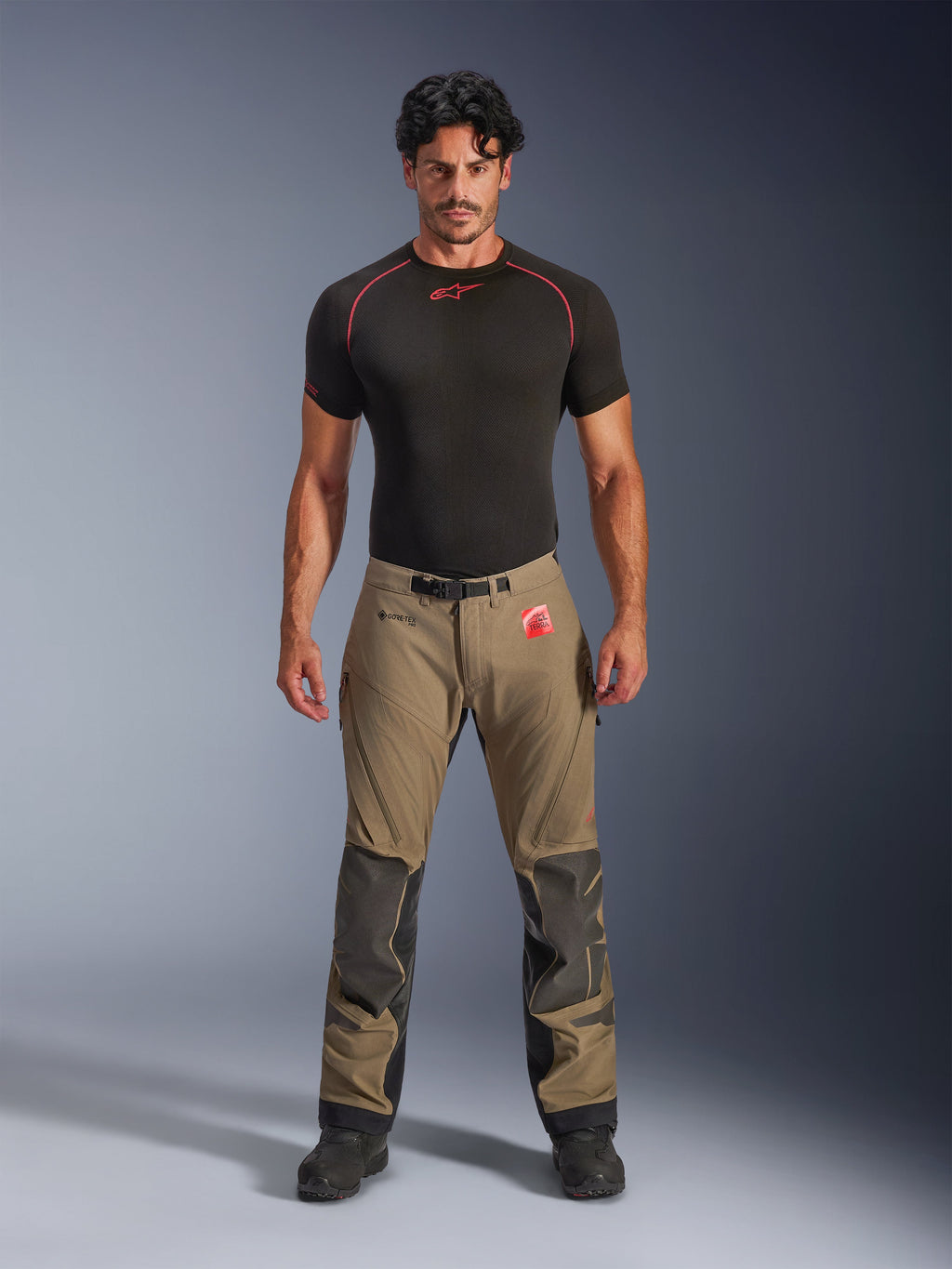Mandlig model iført Alpinestars Nazca 3L GORE-TEX PRO bukser i valnøddebrun og sort, teknisk adventure-køreudstyr med Superfabric-knæforstærkninger og magnetisk taljespænde, kombineret med en sort Alpinestars base layer-trøje, studiebillede set forfra.