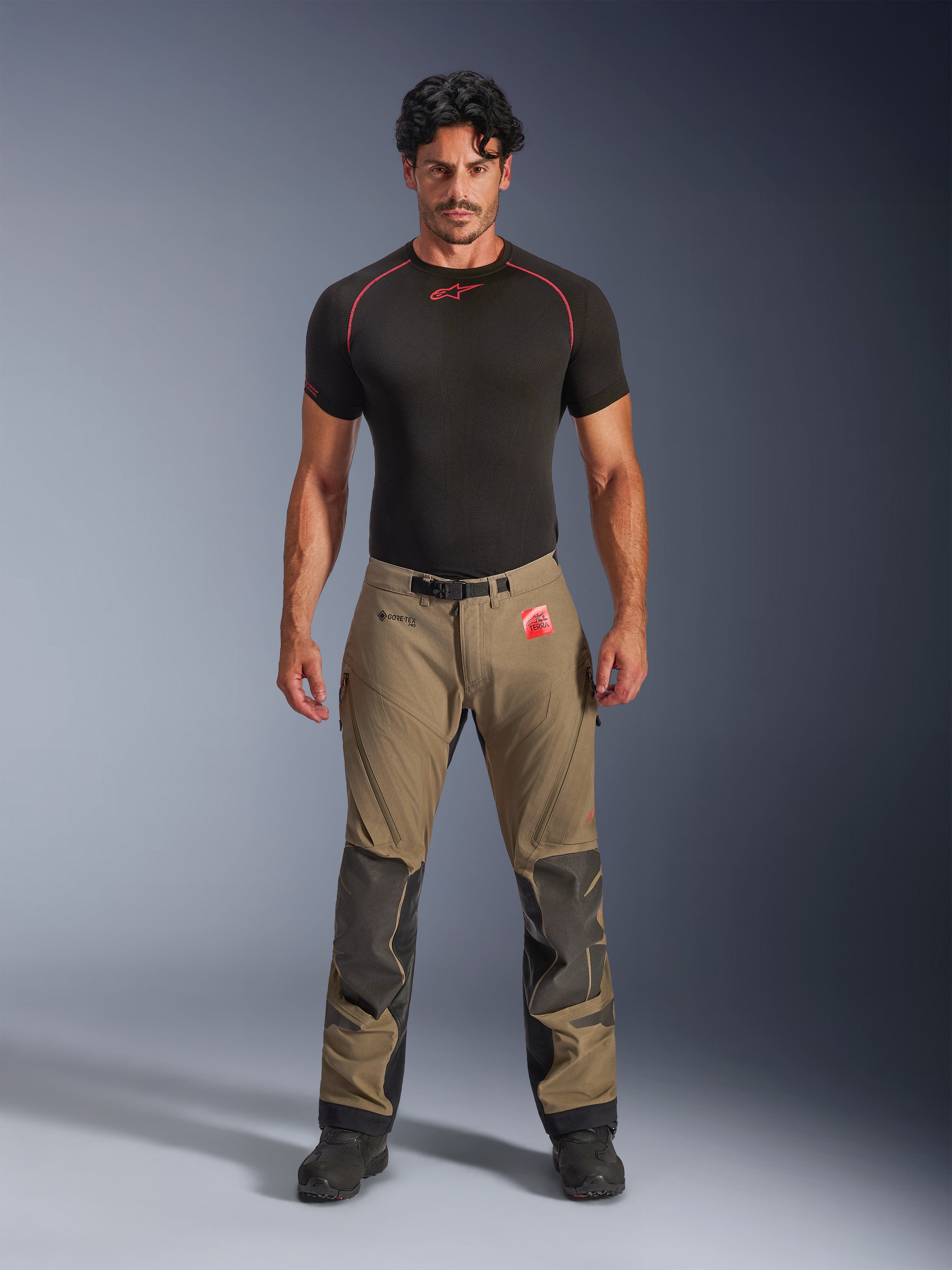 Mandlig model iført Alpinestars Nazca 3L GORE-TEX PRO bukser i valnøddebrun og sort, teknisk adventure-køreudstyr med Superfabric-knæforstærkninger og magnetisk taljespænde, kombineret med en sort Alpinestars base layer-trøje, studiebillede set forfra.