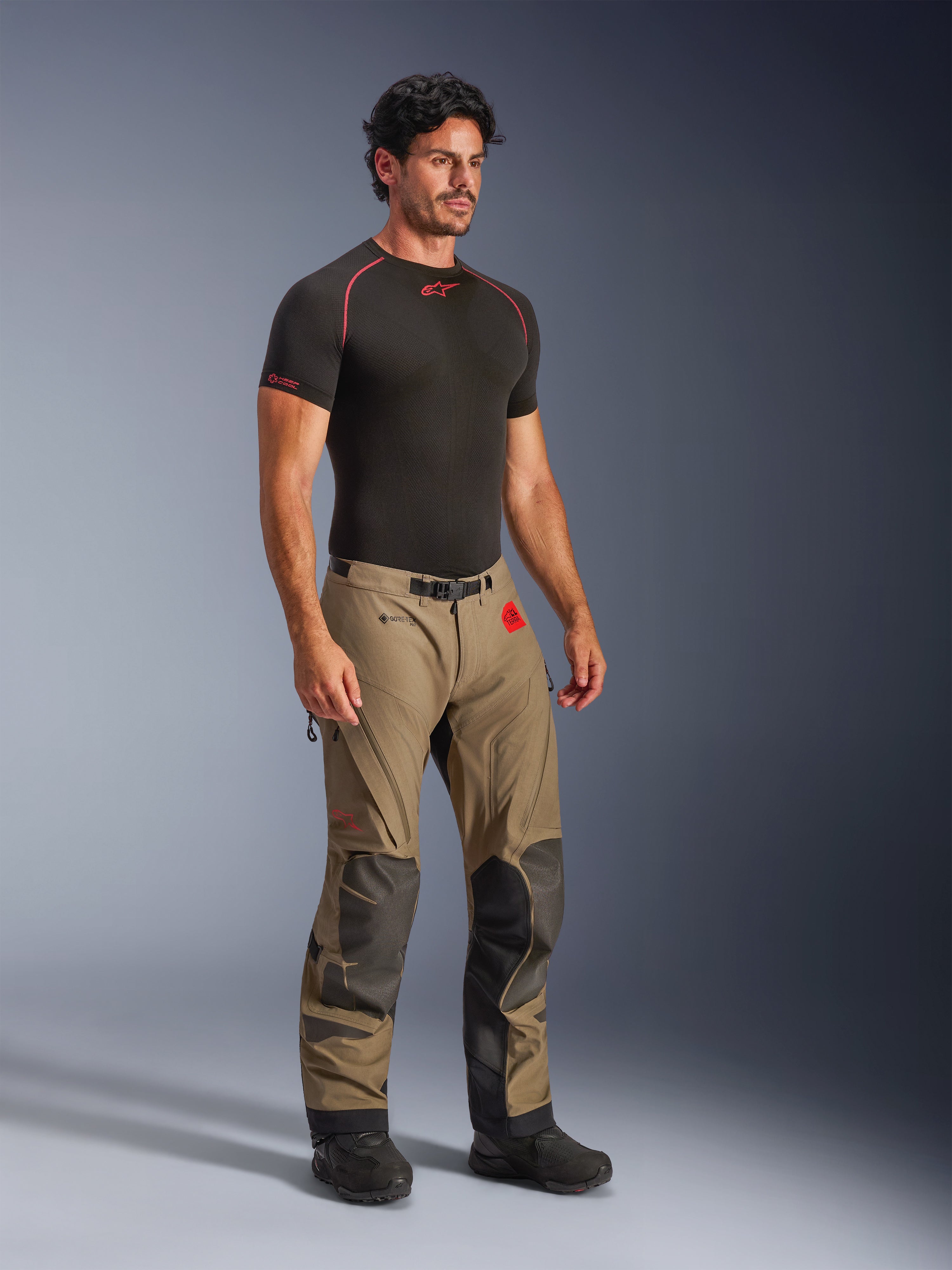 Alpinestars Nazca 3L GORE-TEX PRO bukser i Walnut Black, adventure-motorcykelbukser med sorte Superfabric-forstærkninger på knæene, båret af en model med et sort kompressions-baselayer, bælte med magnetisk spænde og rød All Terra-branding.