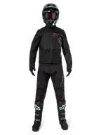 Alpinestars Pro-Dura jakke, sort off-road enduro-jakke, vist på en model med et komplet udstyrssæt inklusiv sort hjelm med røde goggles, sorte bukser med røde detaljer, sort-hvide støvler og sorte handsker, rally-inspireret robust design til terrænkørsel
