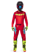 Motocross rider wearing complete racing gear, Red jersey with yellow fluo astars logo, Alpinestars 2025 Supertech Maker Pants in Bright Red and Yellow Fluo, featuring blue logos on thighs, yellow fluo gloves and boots, black helmet with blue goggles, coordinated pro-level motocross outfit