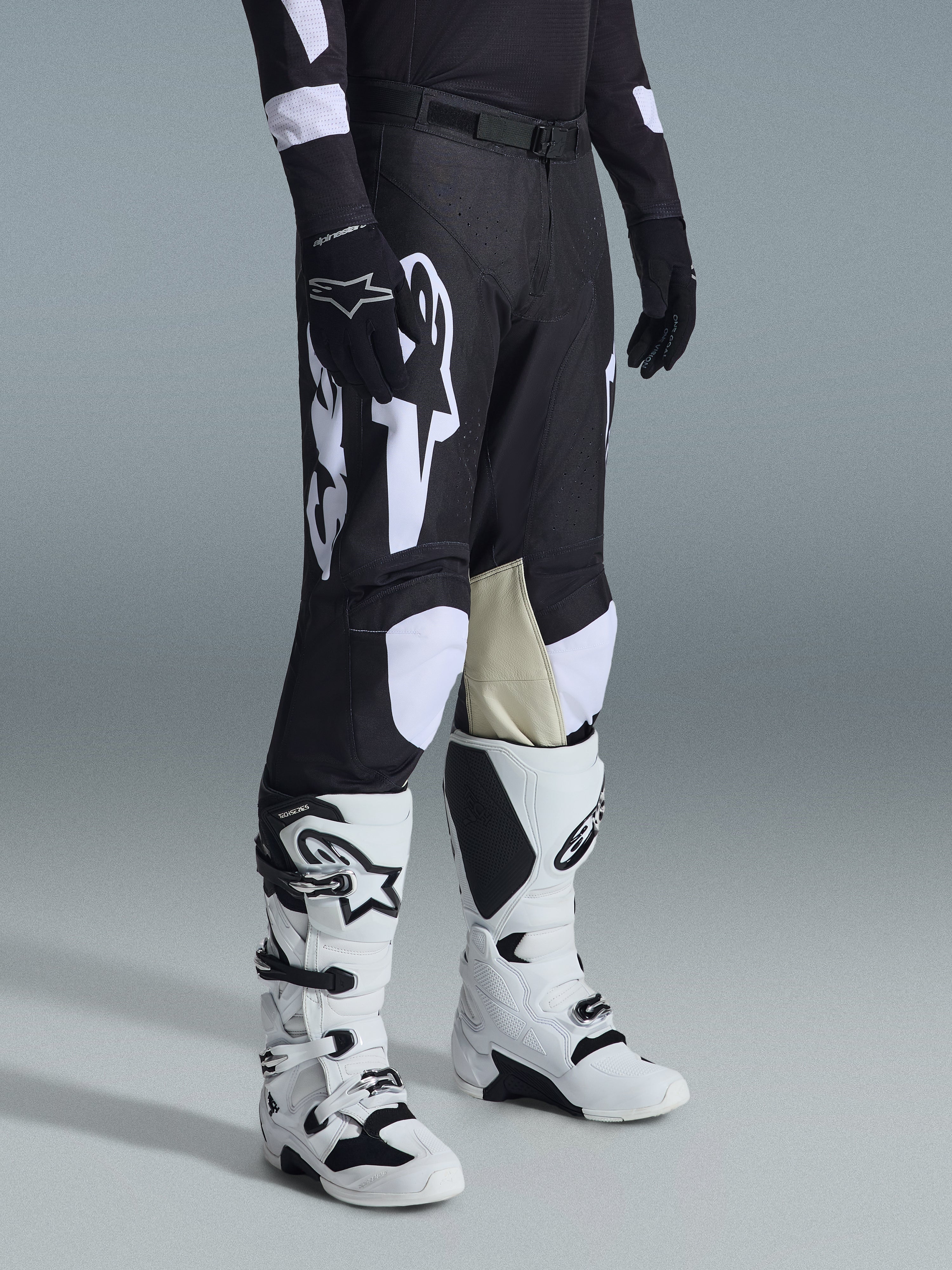 Alpinestars 2026 Racer Air Riway Pants, Black and White motocross pants with large white logo graphics, beige leather inner knee panels, worn with white and black off-road boots and black gloves in a studio setting.