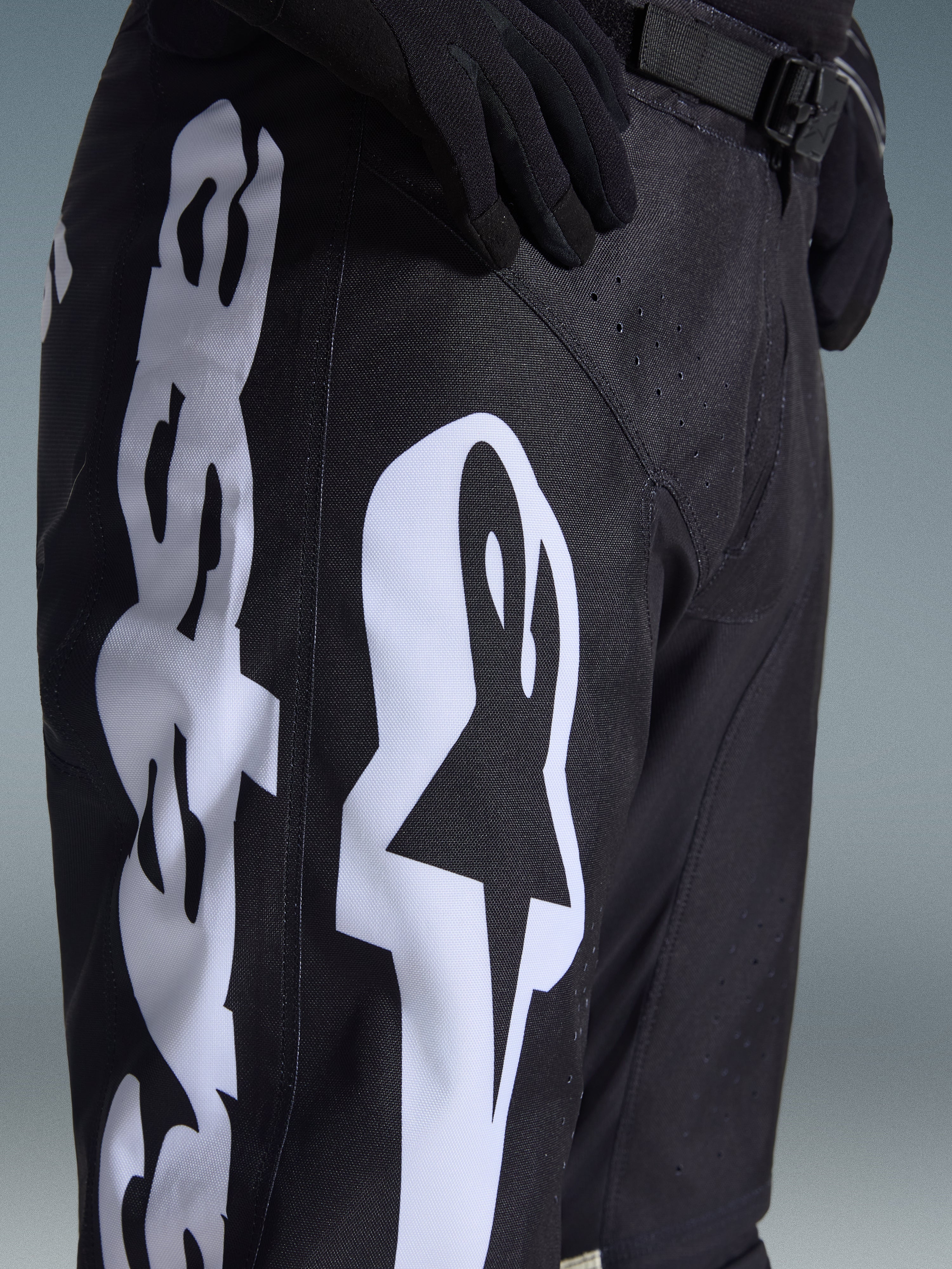 Alpinestars 2026 Racer Air Riway Pants, black motocross pants with bold white Alpinestars logo on the thigh, close-up showing technical fabric and adjustable waist strap, worn with black gloves for off-road riding
