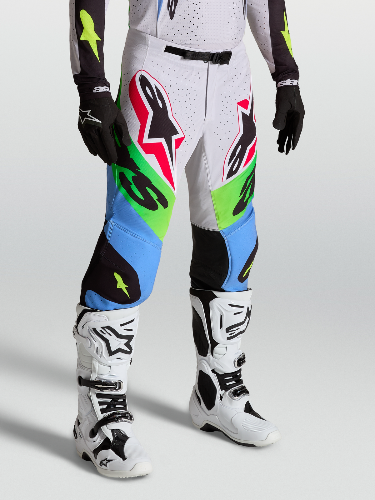 Alpinestars Supertech Dyna Bukser, professionelle motocrossbukser, grønne og blå med hvide detaljer, båret med hvide støvler og sorte handsker til terrænkørsel.