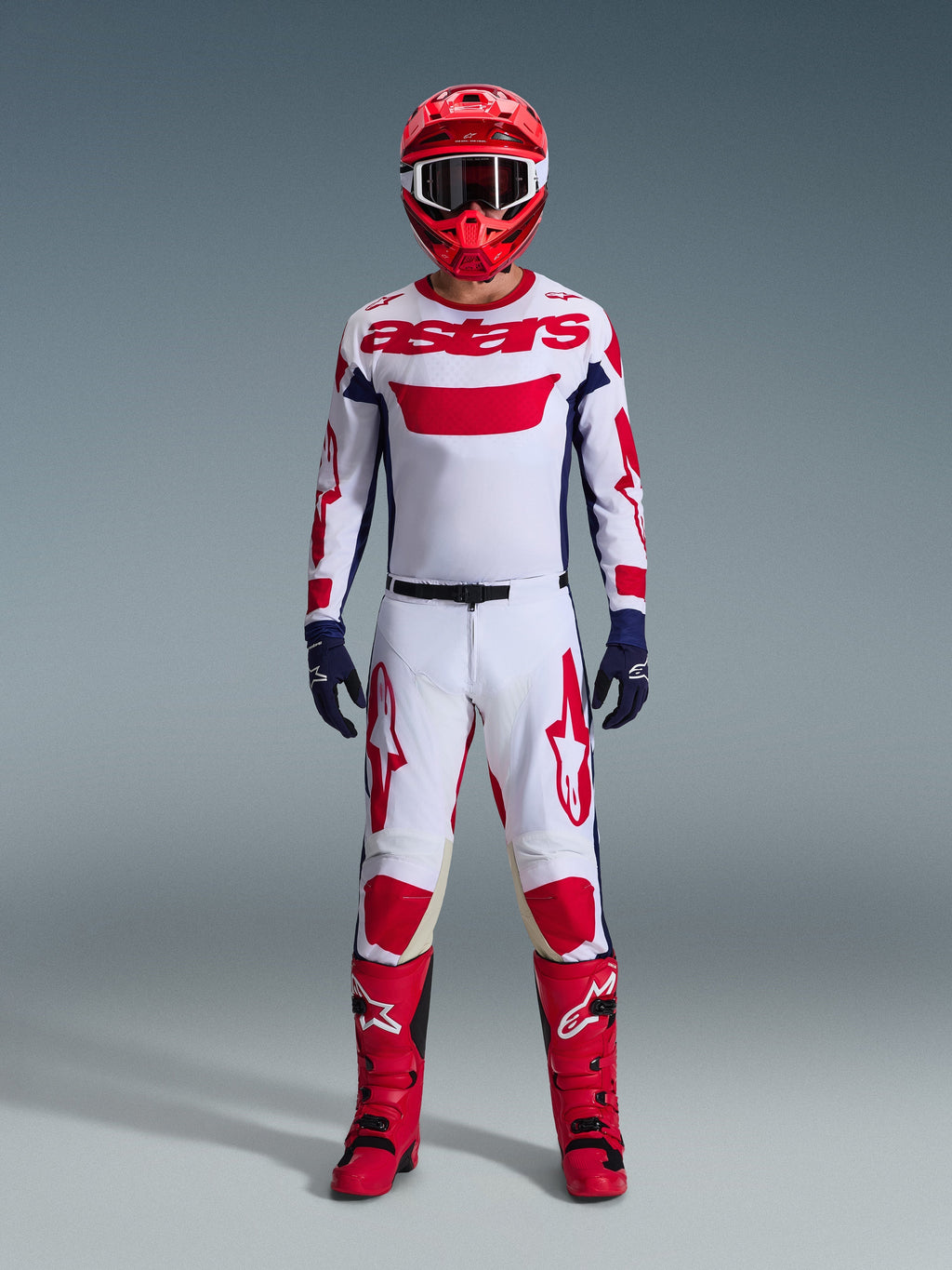 Motocrosskører iført komplet racerudstyr, hvid jersey med rødt Alpinestars-branding og blå detaljer, Alpinestars 2026 Racer Riway bukser i hvid, blå og rød, rød hjelm med matchende goggles, røde og sorte støvler, mørkeblå handsker, koordineret outfit til off-road racing