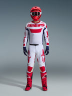 Motocrosskører iført komplet racerudstyr, hvid jersey med rødt Alpinestars-branding og blå detaljer, Alpinestars 2026 Racer Riway bukser i hvid, blå og rød, rød hjelm med matchende goggles, røde og sorte støvler, mørkeblå handsker, koordineret outfit til off-road racing