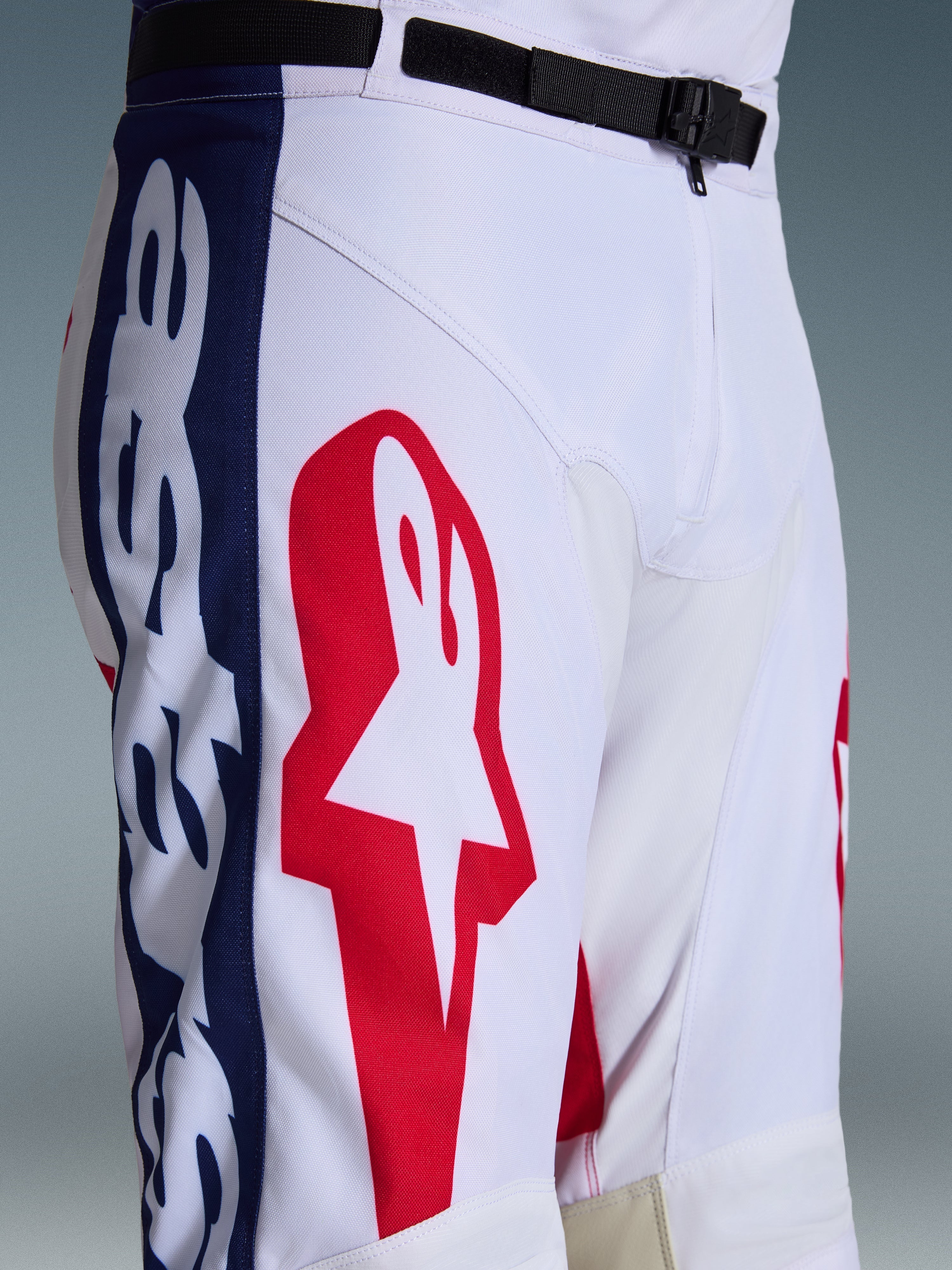 Alpinestars 2026 Racer Riway bukser, motocross-bukser, hvid, blå og rød, nærbillede af øverste del af ben og talje med stort rødt stjernelogo på hvidt polyfabric, blå sidepaneler med branding, sort justerbart taljebælte med SX2 magnetspænde, sporty off-road racerdesign