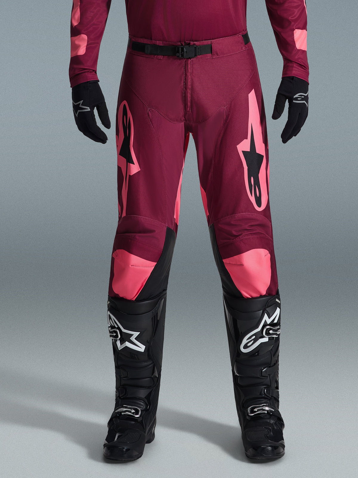 Alpinestars 2026 Racer Riway bukser, motocrossbukser, Dark Red Coral, båret med sorte handsker og støvler, med store koralfarvede Alpinestars-logoer på lårene, sort taljespænde og forbuet bendesign til off-road racing