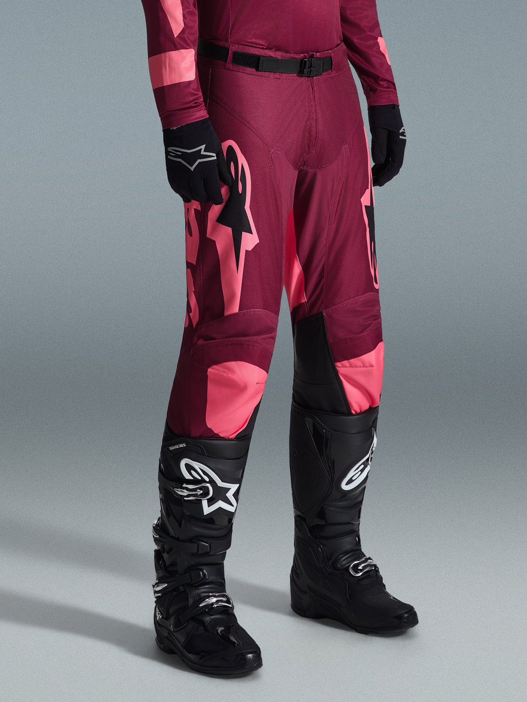 Alpinestars 2026 Racer Riway bukser, motocrossbukser, Dark Red Coral, med store lyserøde stjernelogoer og sorte indvendige knæpaneler, båret med sorte motocrossstøvler og sorte handsker, atletisk racerpasform til off-road kørsel