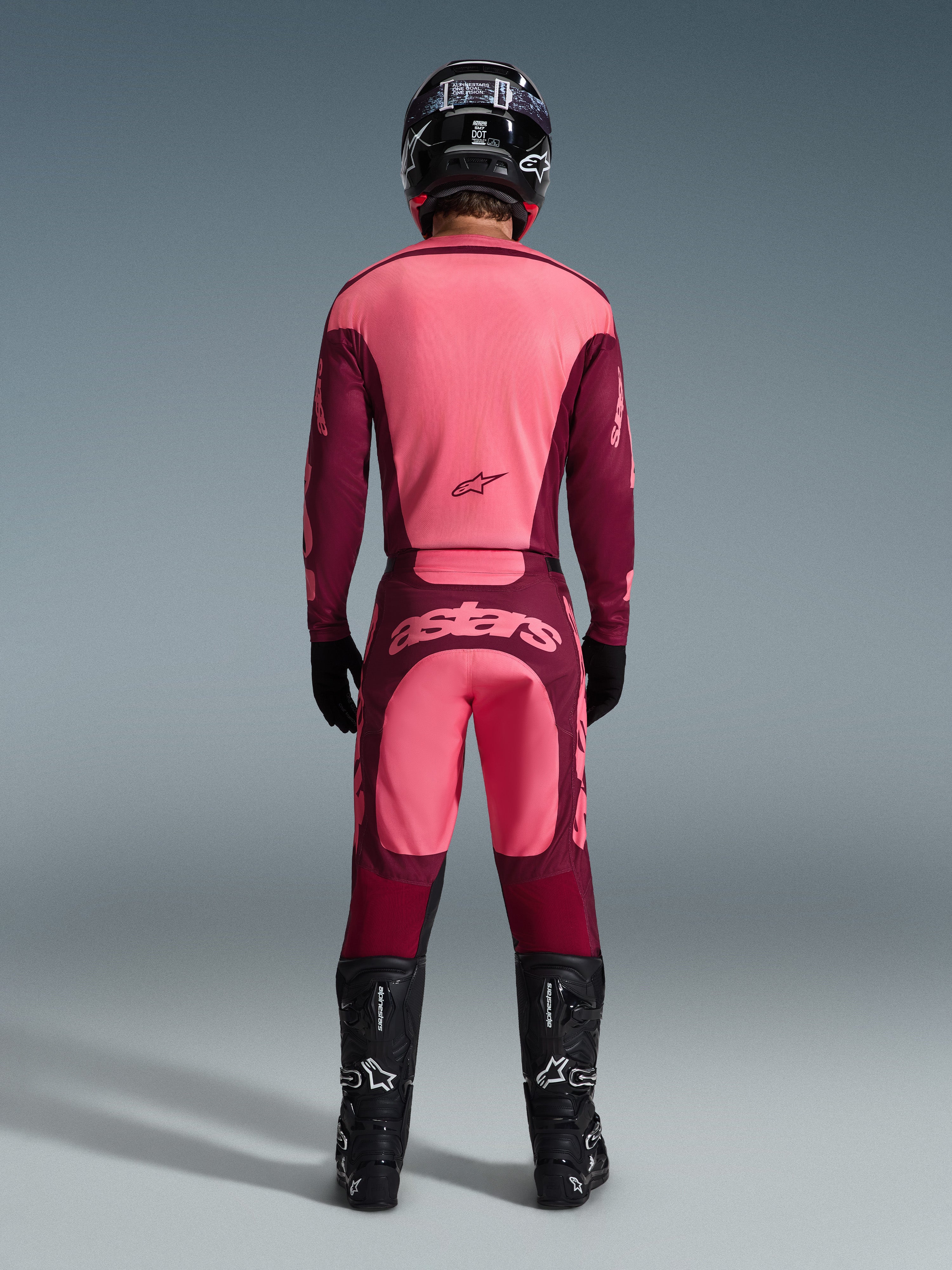 Motocross-rytter set bagfra iført Alpinestars 2026 Racer Riway bukser i Dark Red Coral, matchende trøje, sort hjelm, sorte handsker og sorte Alpinestars støvler, med et markant 'astars'-logo på bagdelen og et tofarvet rødt design.