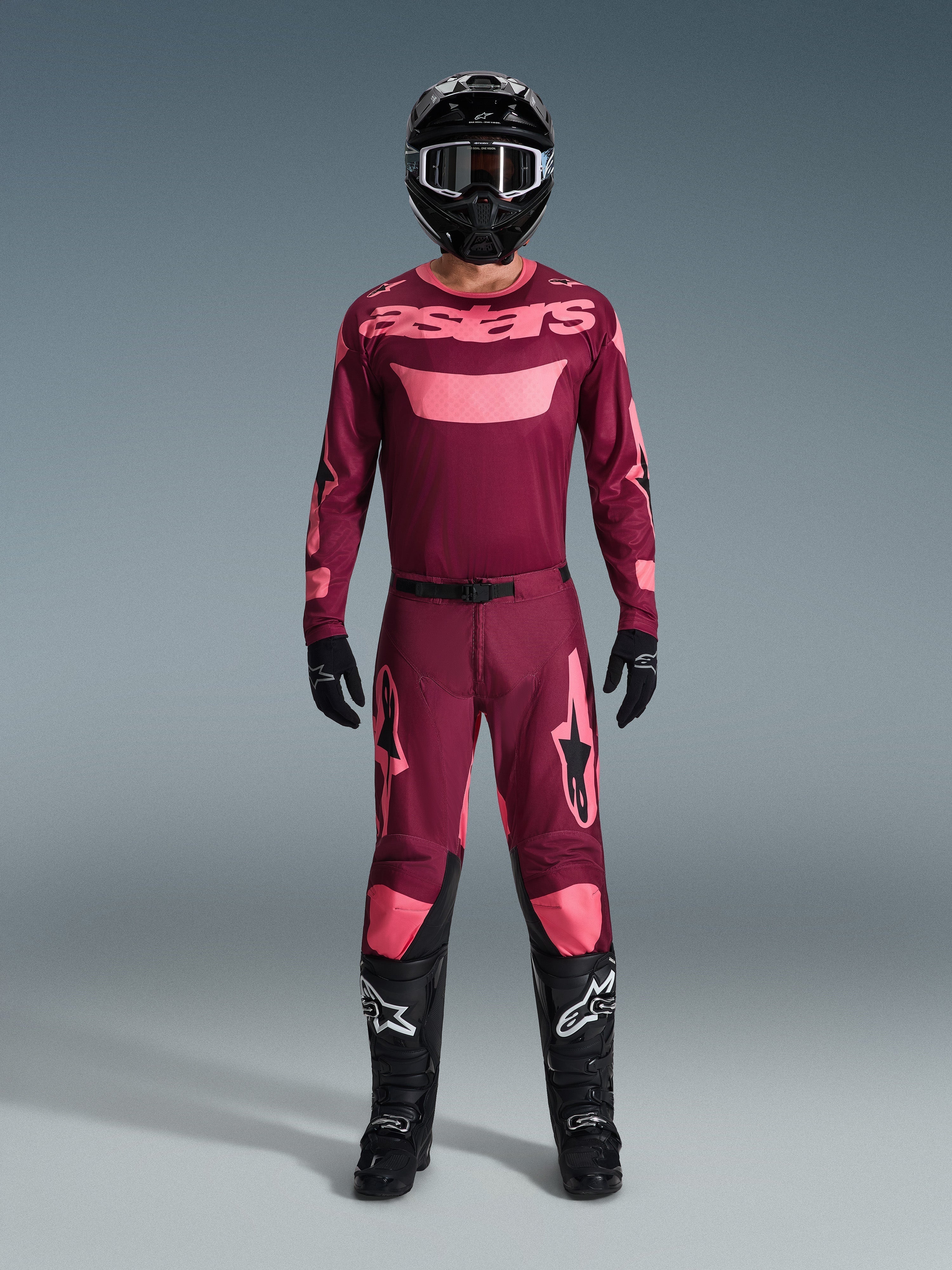 Alpinestars motocross-kører i komplet racerudstyr, Dark Red Coral trøje med pink detaljer, matchende Racer Riway bukser, sort hjelm med hvide detaljer, sorte støvler med hvide logoer, sorte handsker, koordineret outfit til off-road kørsel