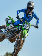 Motocross rider mid-jump on a green motorcycle, wearing Alpinestars 2025 Racer Veil Pants in Blue and Black, matching blue jersey with white chest logo, white and black boots, and black helmet, high-performance off-road gear for racing.