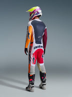 Motocrosskører set bagfra iført komplet racerudstyr, Alpinestars 2026 Racer Portl bukser i rød, lysegrå og mørkegrå, matchende trøje med orange og bordeauxrøde ærmer, flerfarvet grafisk hjelm, grå og sorte off-road støvler med lyserøde detaljer, professionelt motocross-outfit