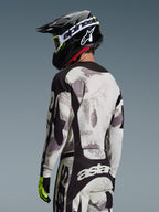 Motocrosskører set bagfra iført 2026 Racer Lahnd-trøje i Sand Camo, grå og hvid abstrakt mønstret trøje med sorte detaljer, kombineret med en sort Alpinestars-hjelm, hvide goggles og neongule handsker, der viser rygprofilen og performance-pasform til off-road racing.