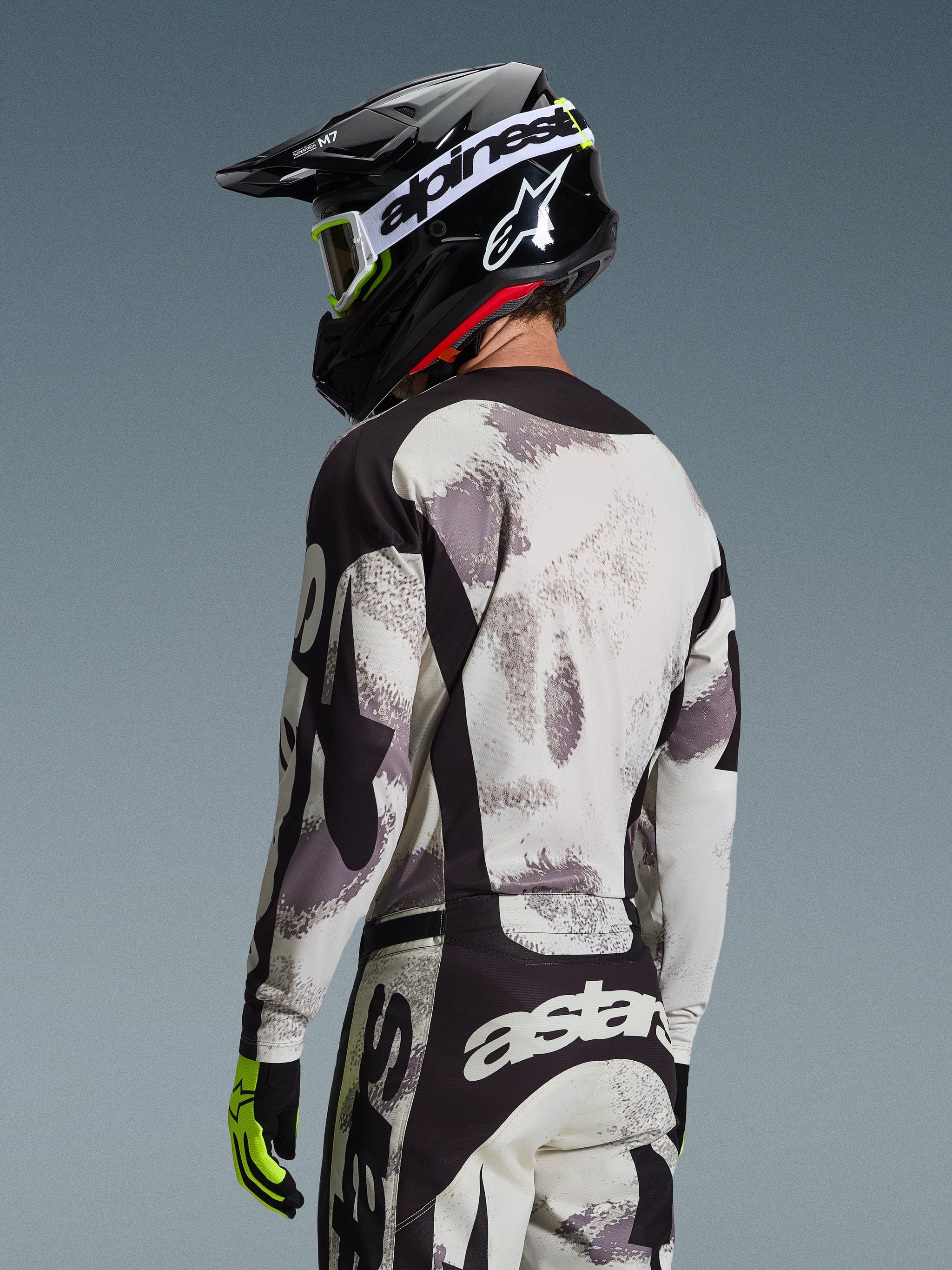 Motocross rider from behind wearing the 2026 Racer Lahnd Jersey in Sand Camo, gray and white abstract patterned jersey with black accents, paired with a black Alpinestars helmet, white goggles, and neon yellow gloves, showcasing the back profile and performance fit for off-road racing.