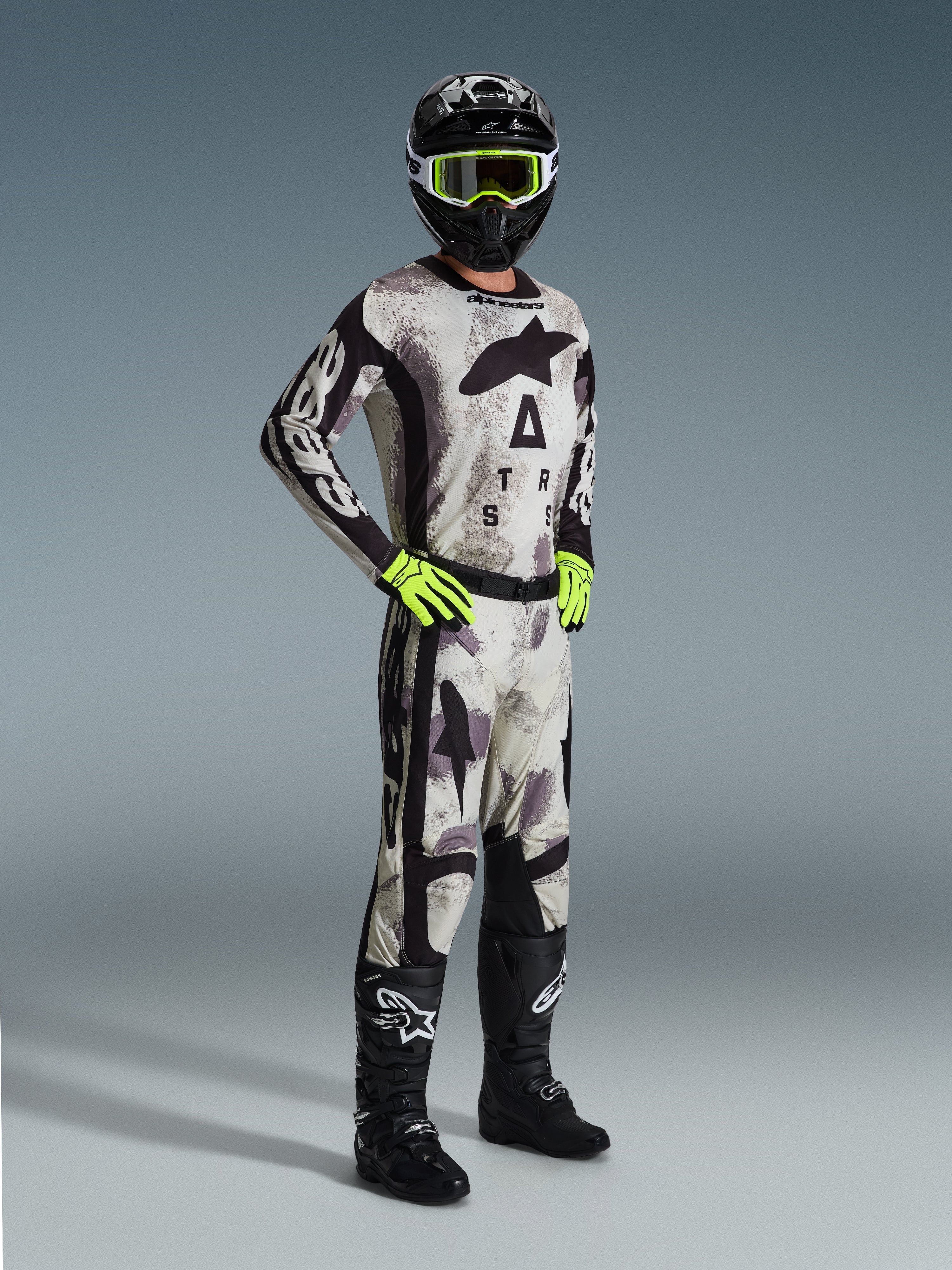 Motocross rider wearing the 2026 Racer Lahnd Jersey in Sand Camo, gray and black camouflage pattern with Alpinestars branding, matching motocross pants, neon yellow gloves, black helmet with yellow goggles, and black boots, complete professional off-road racing outfit.