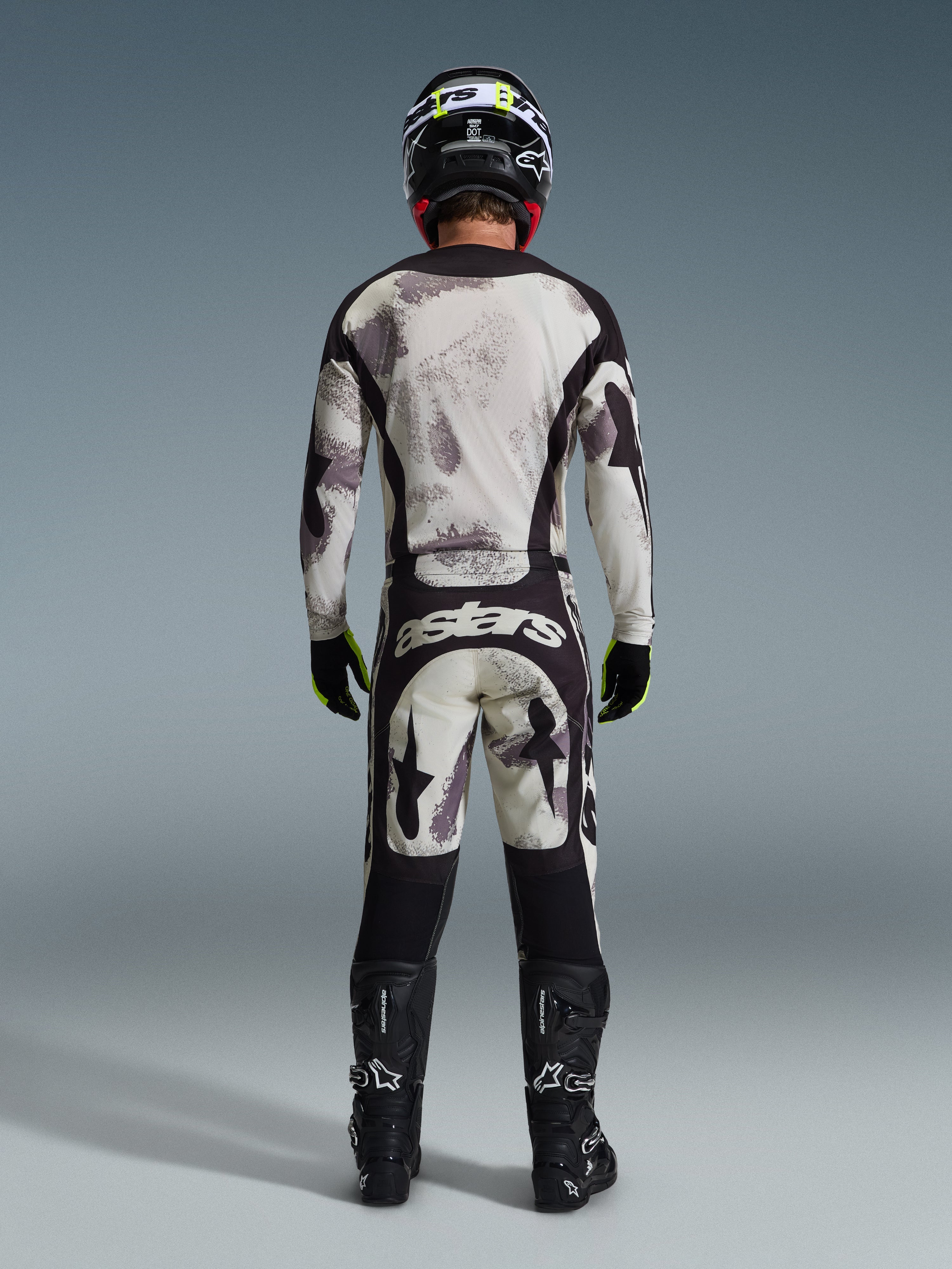 Motocross rider from behind wearing the 2026 Racer Lahnd Jersey and matching pants in Sand Camo, featuring a cream and grey abstract pattern with black panels, 'astars' branding on the pants, paired with black Alpinestars boots and a black helmet for a complete off-road racing look.