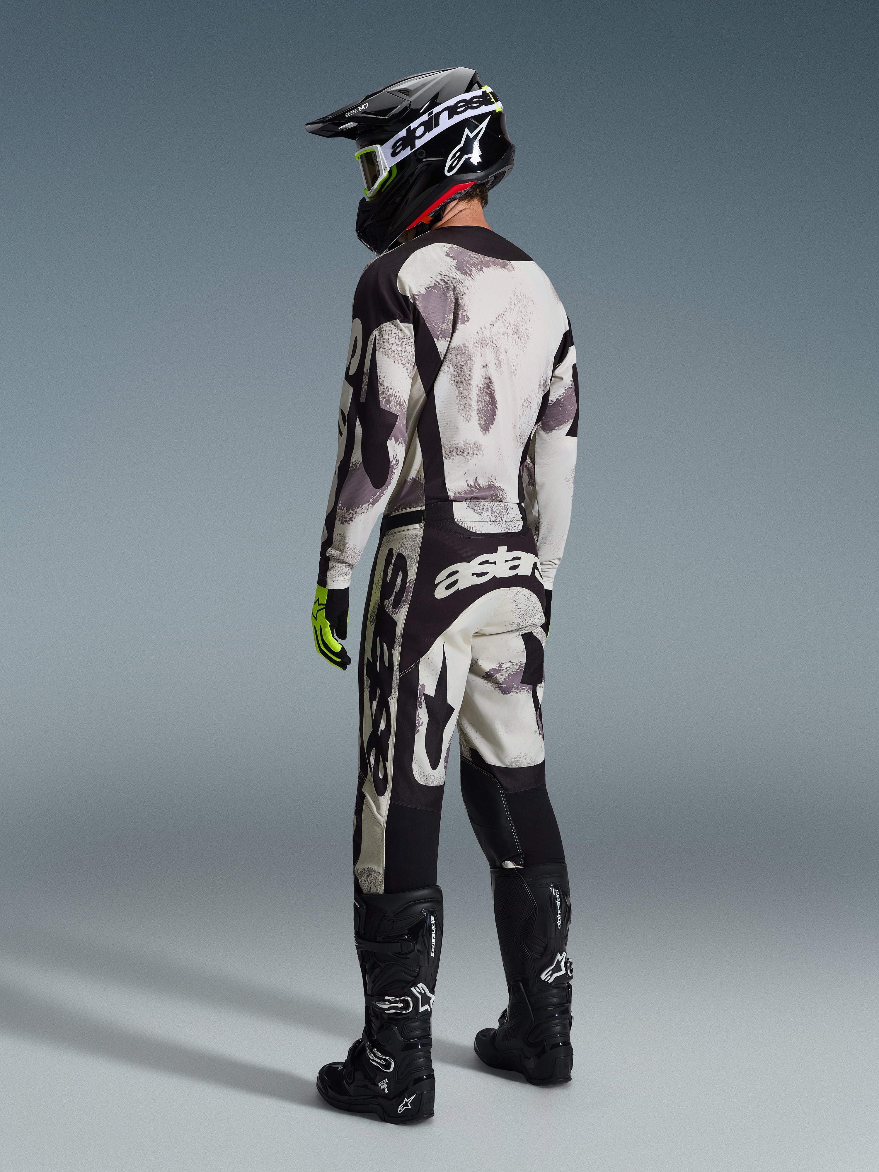 Motocross rider wearing complete racing gear, 2026 Racer Lahnd Jersey and matching pants in Sand Camo, featuring a gray and black camouflage pattern with bold Alpinestars logos, black helmet, black boots, and neon yellow gloves, rear profile view.